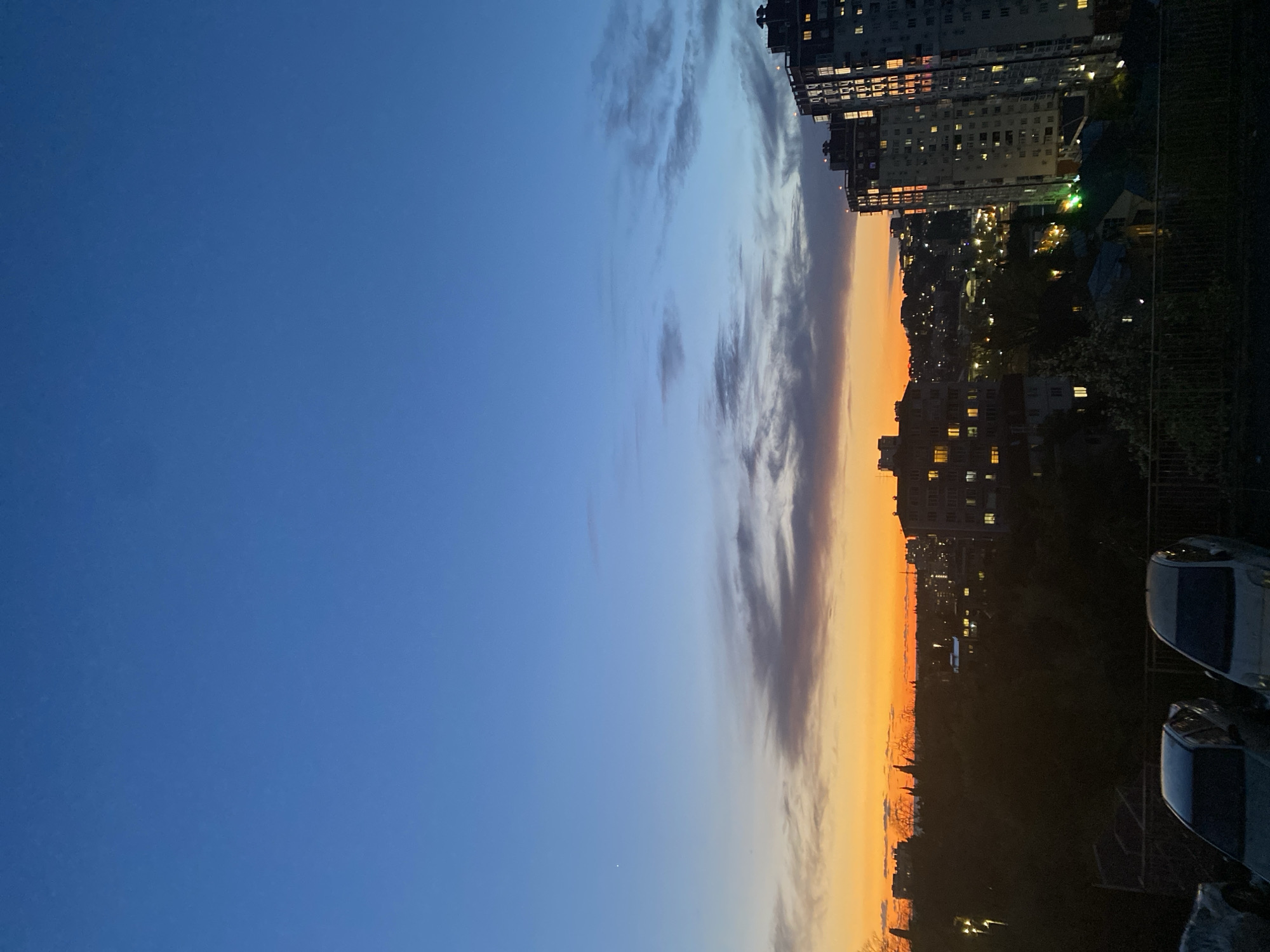 The image shows a cityscape at dusk, likely in Sochi, Russia, given the architecture and general appearance. The sky is transitioning from a clear blue on the left to an orange-yellow hue near the horizon in the center. Several elongated clouds are visible spanning vertically across the image in the transition zone of the sky. High-rise buildings with illuminated windows stand out against the darkening sky on the right side. A lower level with trees and foliage is present, with scattered lights suggesting buildings and street lamps below. In the bottom right corner, two cars are partially visible.