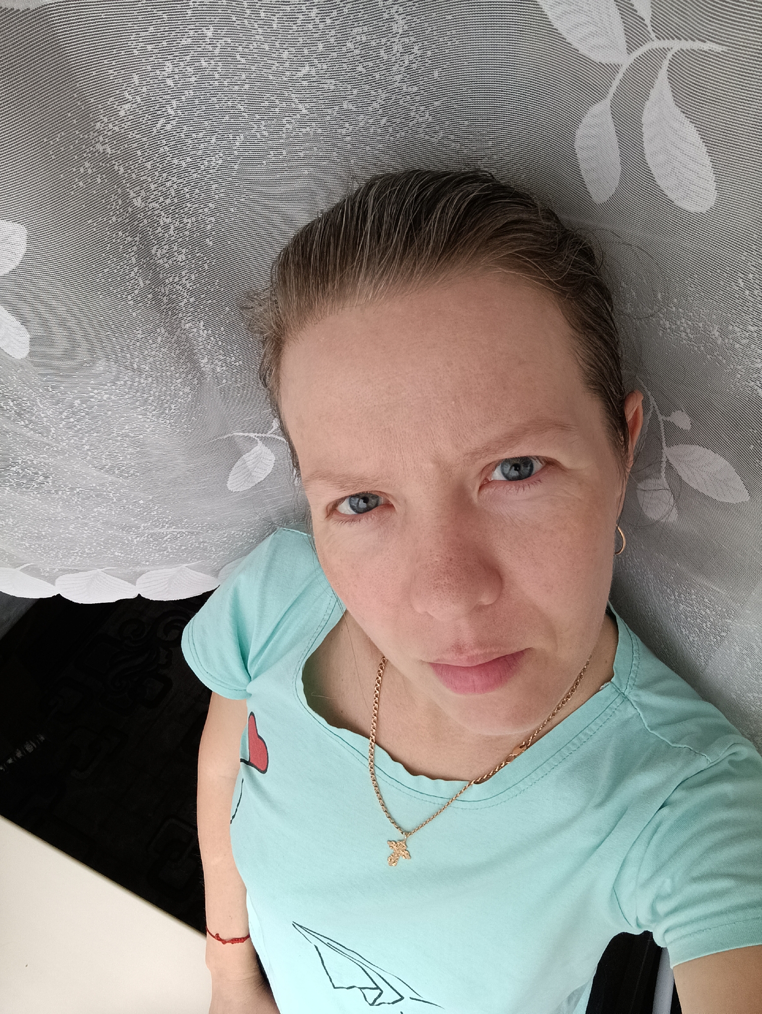 A woman with fair skin, short brown hair with some grey strands, and blue eyes poses for a selfie. She is wearing a light blue t-shirt with a cartoon design and a gold necklace with a cross pendant. She has a small gold hoop earring in her left ear. Behind her is a grey and white patterned curtain. A dark, glossy surface is visible to the left, and part of a white surface is underneath it. A red string bracelet is visible on her left wrist. Based on geotagging and context, the location is determined to be in the city of Chaikovsky, Russia.