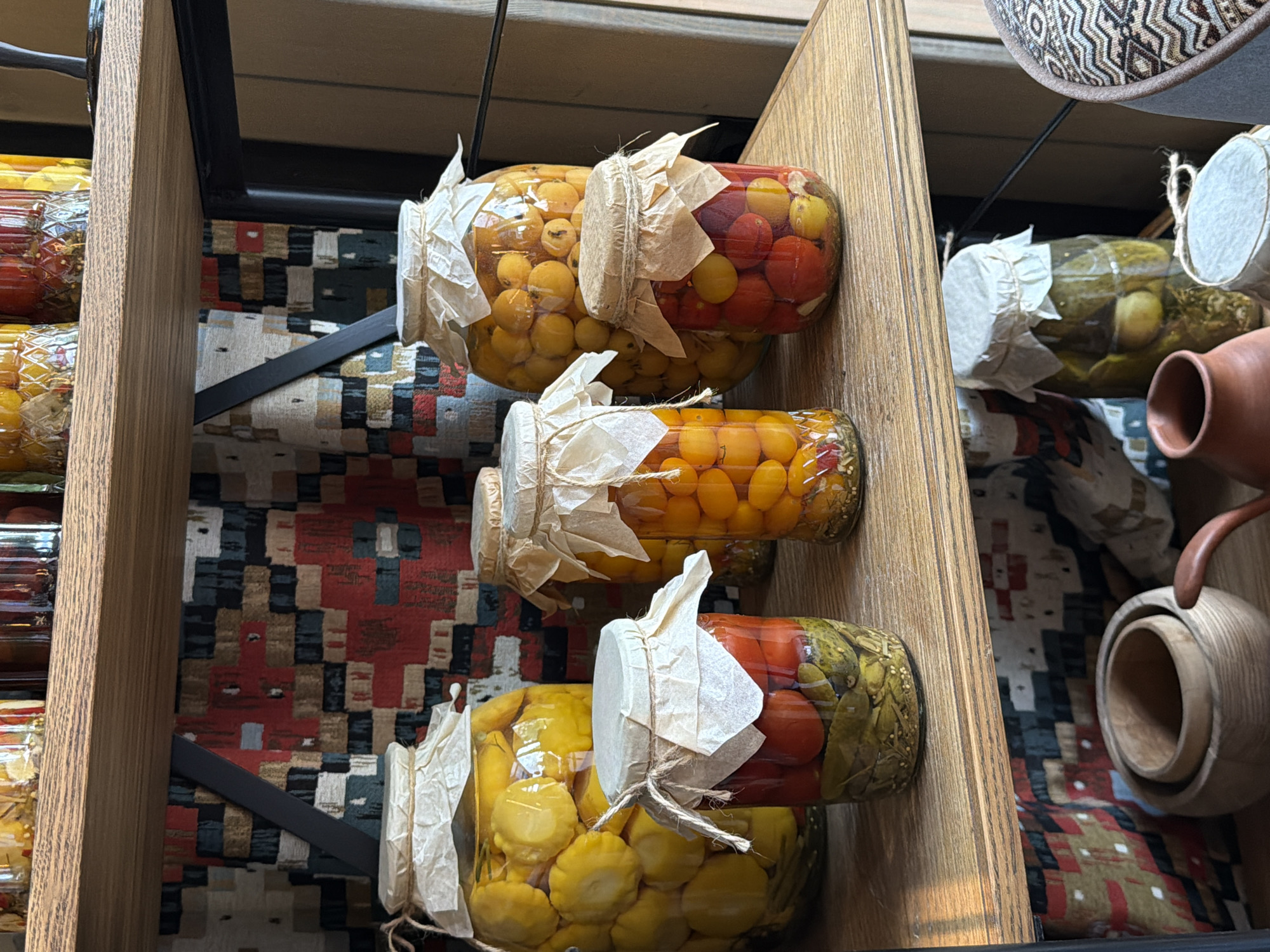 The image shows shelves filled with glass jars containing pickled vegetables. The jars are sealed with paper and twine. The vegetables appear to be tomatoes, yellow squash, and other unknown vegetables. The background consists of a patterned fabric or tapestry. Adjacent to the pickled jars are assorted clay and wooden vessels. The photograph likely captures a scene within a market, deli or specialty food shop in Polivanovo, Russia, where these goods might be sold or displayed.