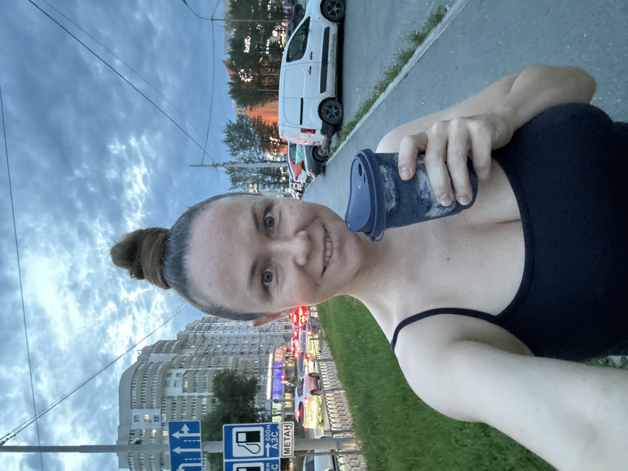 A woman with her hair in a bun holds a dark blue reusable coffee cup in her right hand. She is wearing a black tank top. She is standing on a sidewalk next to a street with traffic. In the background, there are apartment buildings and trees. A street sign with symbols and text, including what appears to be Cyrillic, is visible on the left side of the image. The sky is overcast. The location is likely Yekaterinburg, Russia, based on visual cues.