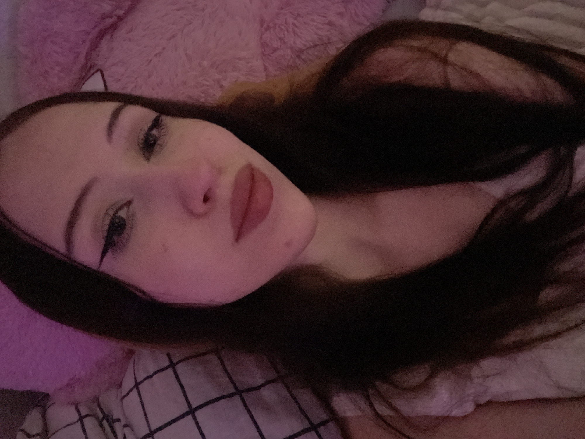 The image shows a close-up of a young woman lying down, with her head and shoulders visible. She has long, dark hair, and is wearing a white top with a textured pattern. She is looking towards the camera, and her makeup includes winged eyeliner and lip gloss. Behind her, there appears to be a pink, fluffy object, possibly a pillow or stuffed animal, and a black and white checkered pattern, which could be bedding. The lighting in the image is dim and tinted with pink, suggesting an indoor setting, possibly a bedroom. There is no indication of the city of Votkinsk, Russia in the image.