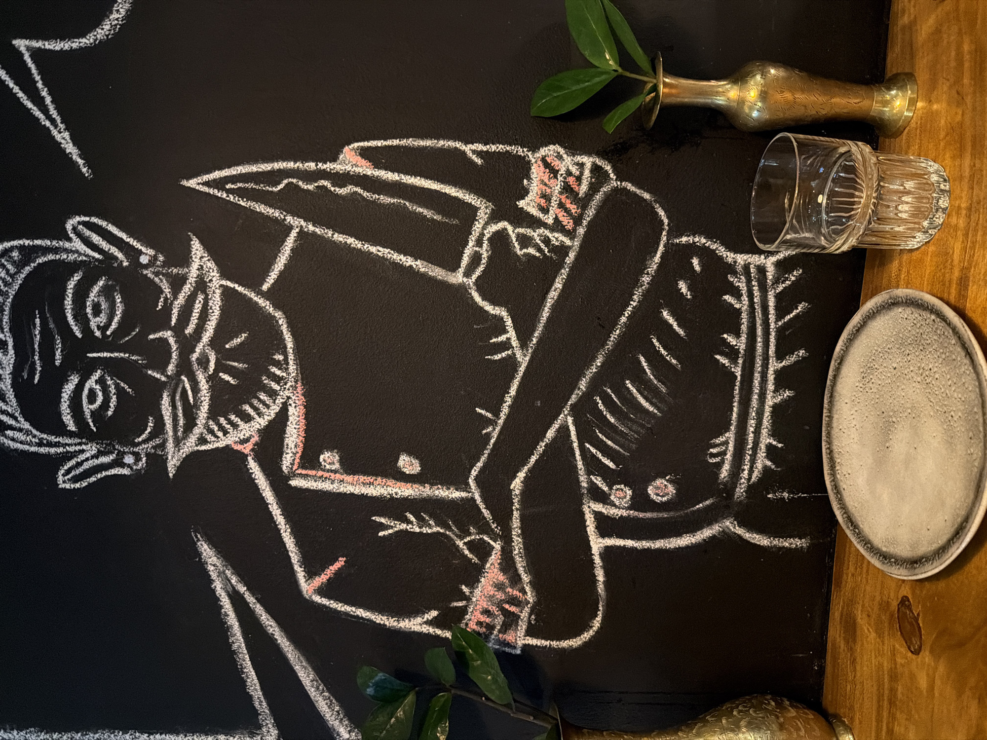 A chalkboard drawing of a man with horns is situated next to various items. A brass vase with greenery, a drinking glass, and a saucer are positioned on a wooden surface. The image was likely taken indoors. The location in the city of Sofia, Bulgaria is not discernable from the image.