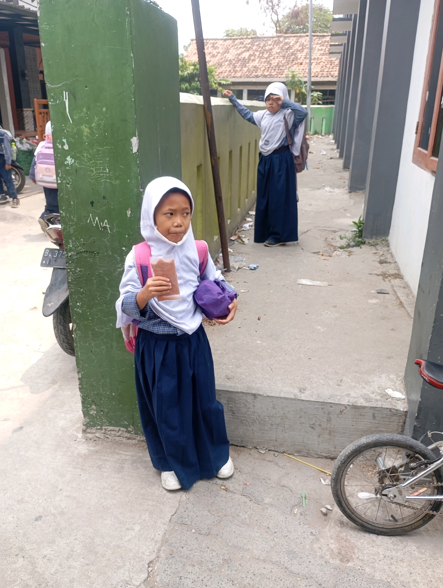 In Babelan, Indonesia, a young girl wearing a white hijab and a blue skirt stands on a concrete pathway. She holds a small, brown packet in her right hand and a purple pouch in her left. Her backpack, a mix of pink and white, is slung over her shoulder. To her left, a green pillar stands prominently. Behind her, another girl in similar attire, also wearing a hijab and blue skirt, is visible further down the path, with a backpack on. A motorcycle is parked to the left of the first girl, partially obscured by the green pillar. The environment appears to be outdoors, with buildings visible in the background, suggesting a school or public area. A bicycle wheel is visible on the right side of the image.