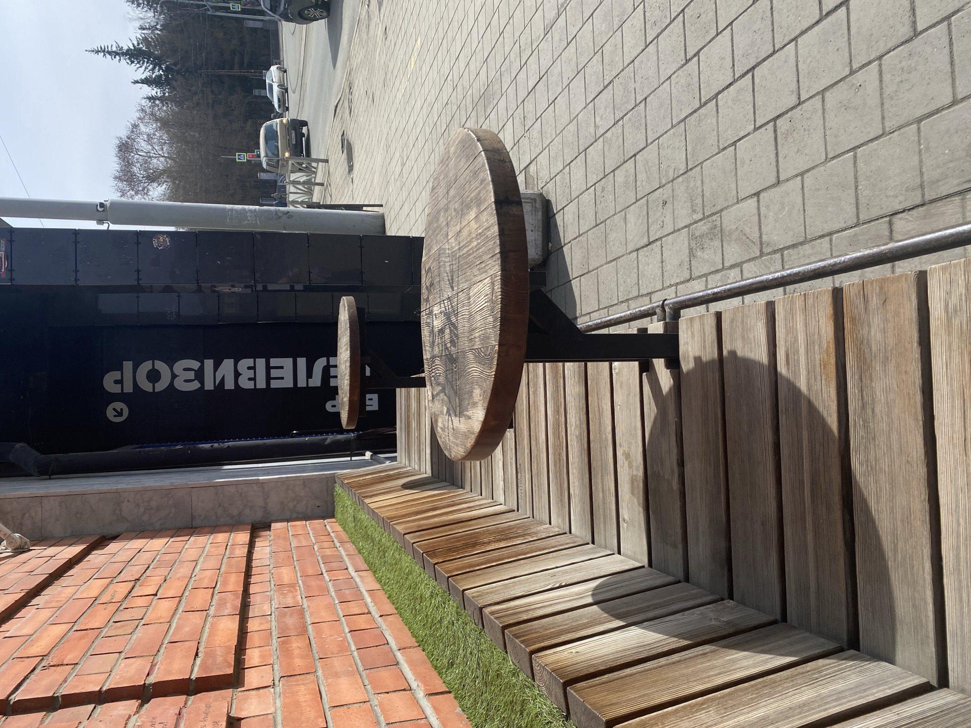 The scene shows an outdoor seating area in Yekaterinburg, Russia. The seating consists of a wooden bench with a low wooden wall behind it. Attached to the wall are two round wooden tables on metal supports. The seating area is next to a brick-paved surface with a strip of artificial turf in between. Behind the seating area is a dark-colored structure with text in Russian. In the background, a street with cars and trees is visible. The sky is overcast.