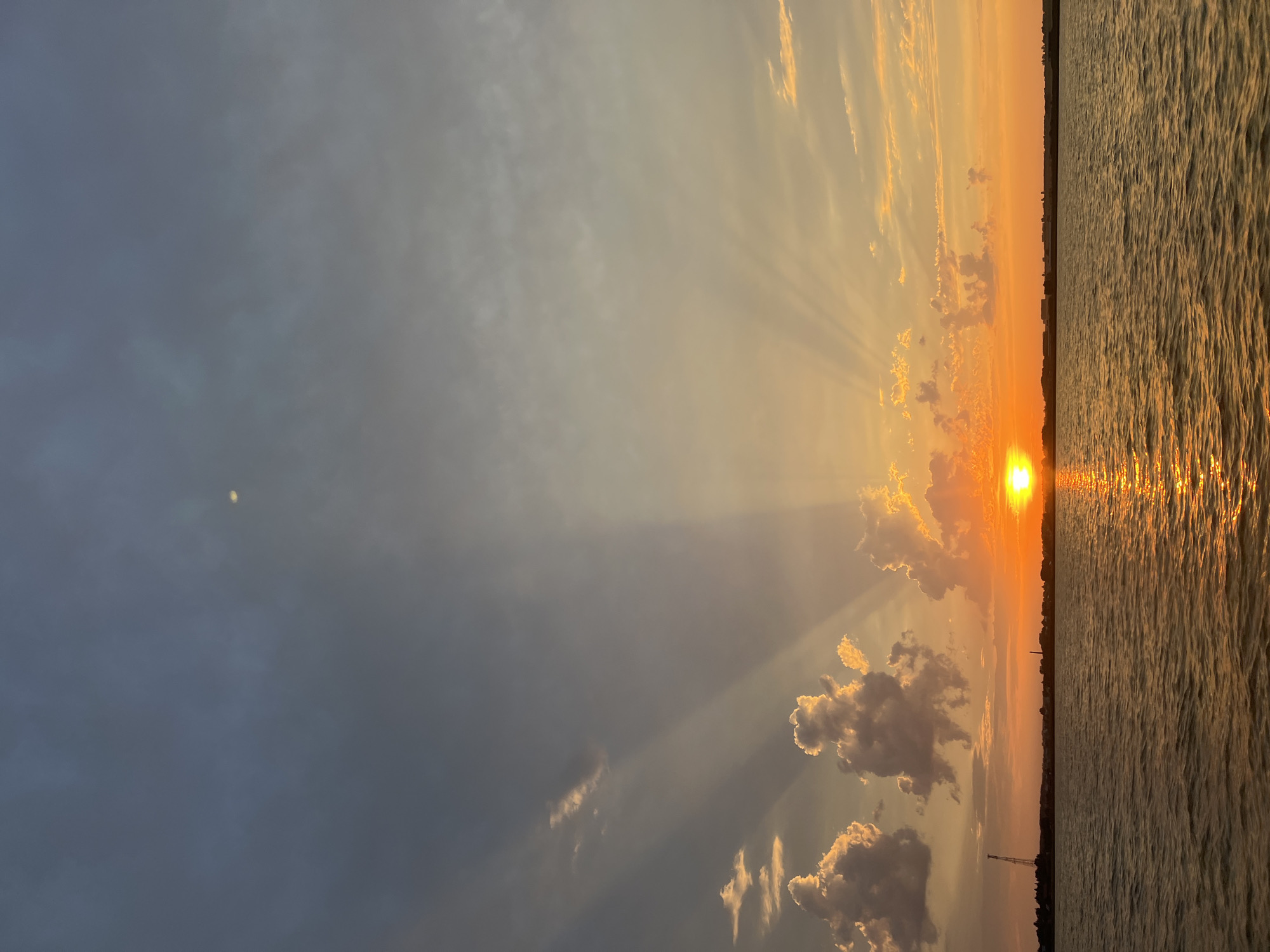 The image depicts a sunset over a body of water. The sun is a bright orange orb positioned on the right side of the horizon, casting brilliant rays upwards into the sky. The sky transitions from a deep blue at the top to warm oranges and yellows near the horizon, interspersed with scattered clouds that are illuminated by the setting sun. The water reflects the colors of the sky and the sun, creating a shimmering surface. On the horizon, there is a faint silhouette of a distant shoreline with some structures, including what appears to be a tall crane on the left side. The overall scene conveys a sense of natural beauty and tranquility. The location of Novopichugovo, Russia is not explicitly visible in the image, but the scene is consistent with a rural or semi-rural setting near a large body of water. There are no people or specific actions captured in the image.