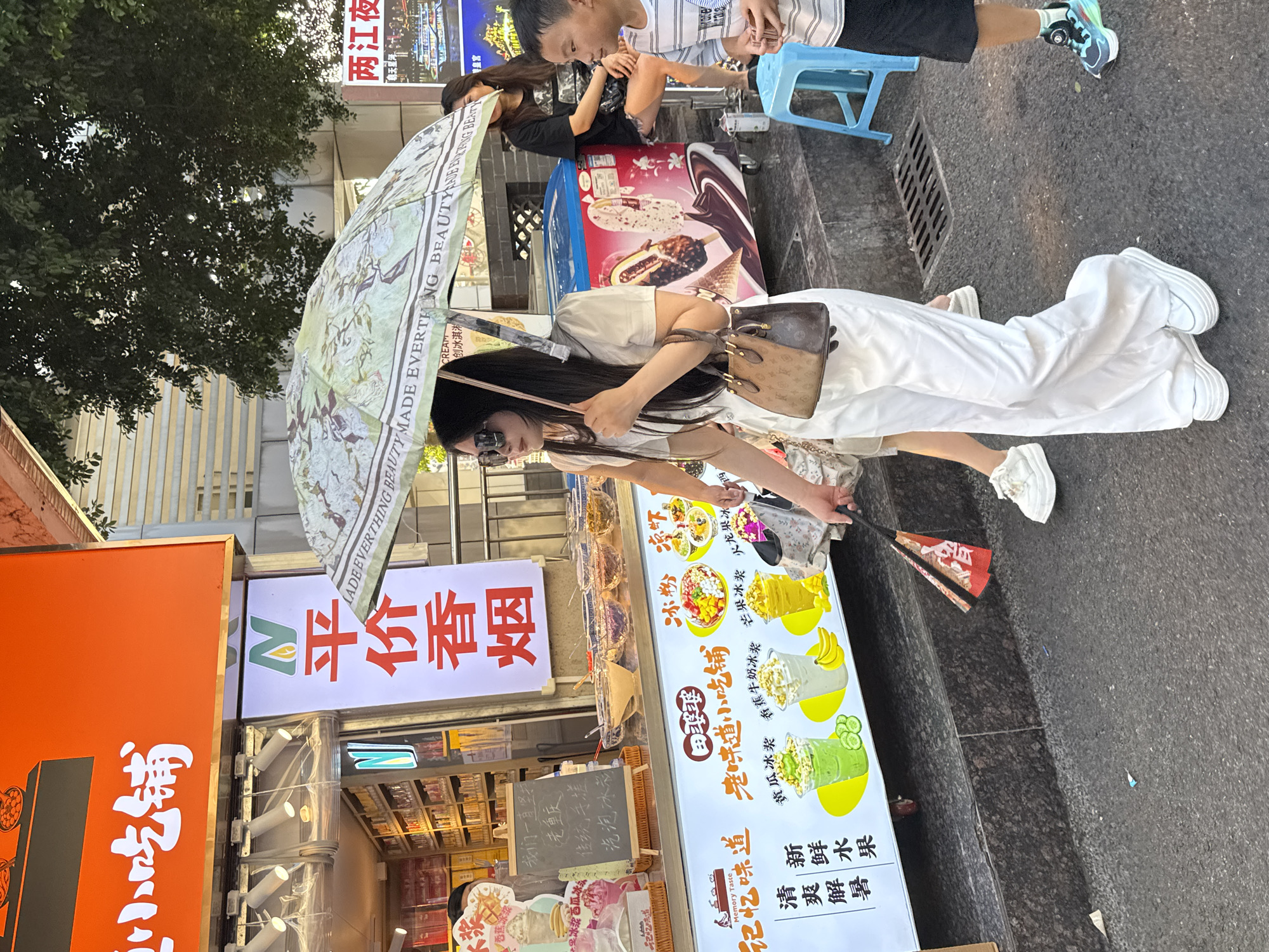 In the Yuzhong District of China, on a city street, a woman walks under a patterned umbrella. She is dressed in loose white pants and a light-colored top, carrying a brown handbag. To her right, a young boy in a striped shirt looks forward, and a woman in a black t-shirt sits on a blue stool. Behind them, a street vendor stall displays various ice cream and dessert items. A large orange sign with white Chinese characters hangs above the stall. The scene is captured in daylight, with dappled sunlight filtering through trees overhead.