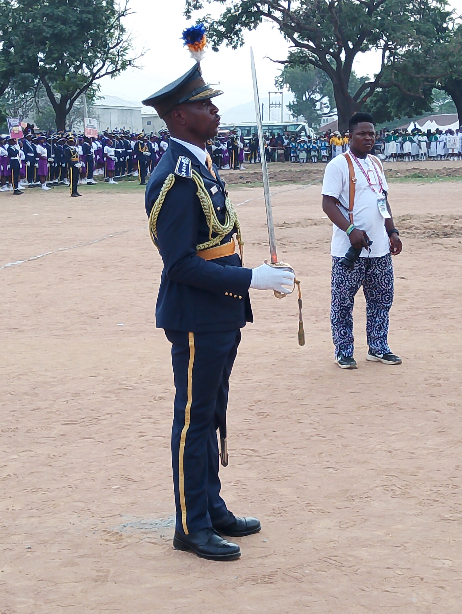The image depicts a man in a blue military uniform with gold braid and a plumed hat, holding a sword. He stands at attention on a dirt field. Behind him and to the right is a man wearing a white t-shirt and patterned blue and white pants, holding a camera with a strap around his neck. In the background, there are groups of people dressed in various uniforms, and trees. The setting is outdoors in Kubwa, Nigeria.