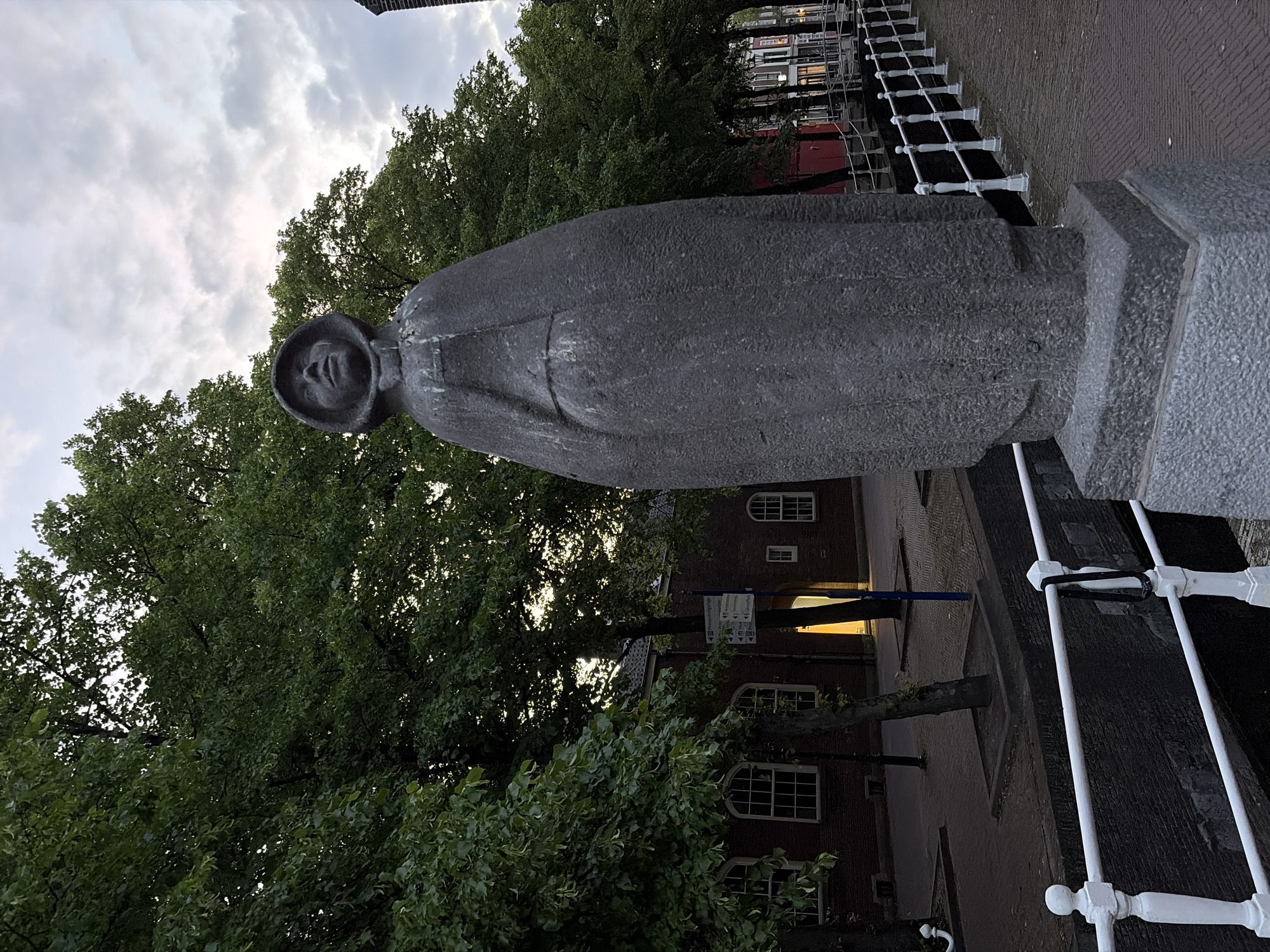 A gray statue of a person in a robe stands beside a bridge in Delft, Netherlands. The statue has a cowl around the head and a simple face. The bridge has white railings. Trees with green leaves are behind the statue and to the left of the frame. Buildings are visible behind the trees. The sky is cloudy.