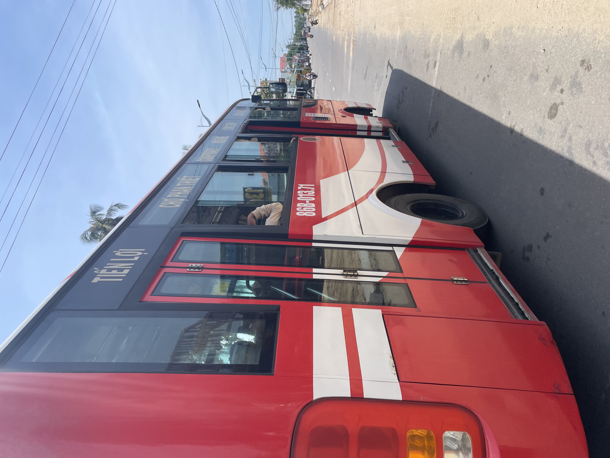 A red and white bus is parked on the right side of a paved road in Phu Thuy Ward, Vietnam. The bus has several windows and two sets of doors on its side. The license plate of the bus is "86B-013.71". Text above the windows on the bus reads "TIEN LOI" and "CHỢ PHAN THIẾT". Other text is also visible but unreadable. The bus is casting a shadow on the road. In the background, there are buildings, trees, and a clear blue sky with some power lines. Several motorcycles and other vehicles are visible on the road in the distance. The environment appears to be a street with some construction or unpaved areas along the side of the road.