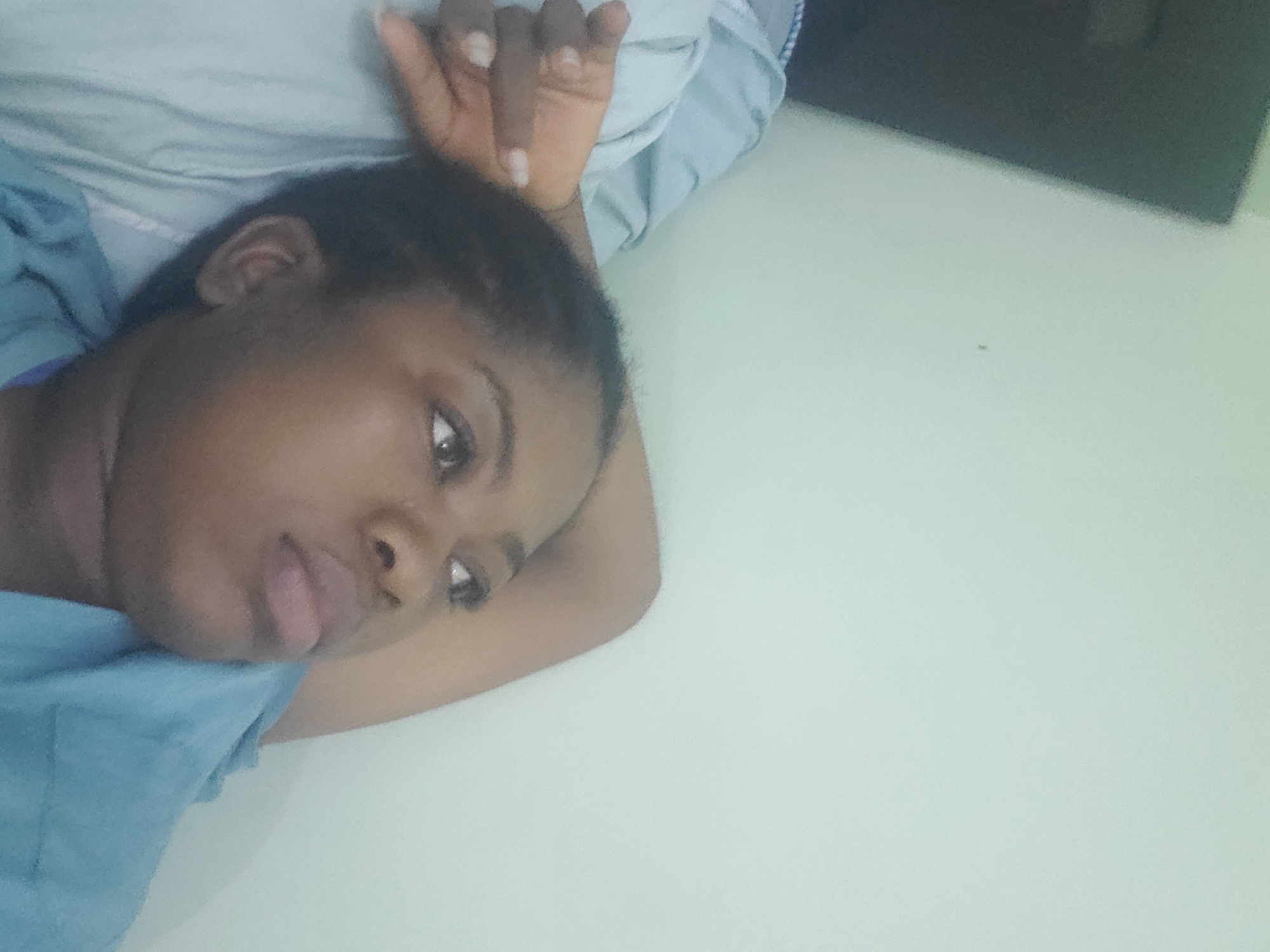 A medium shot captured from a high angle shows a young Black woman lying down, looking upwards and to the right. She has dark skin, dark hair pulled back, and is wearing a light blue garment. Her right arm is bent at the elbow and resting behind her head. Her left hand is resting on her left bicep, with fingers curled. The background is a plain, light-colored wall. The overall scene suggests a relaxed or contemplative moment. The image does not contain specific information to determine its location within Barden Yana, Nigeria.