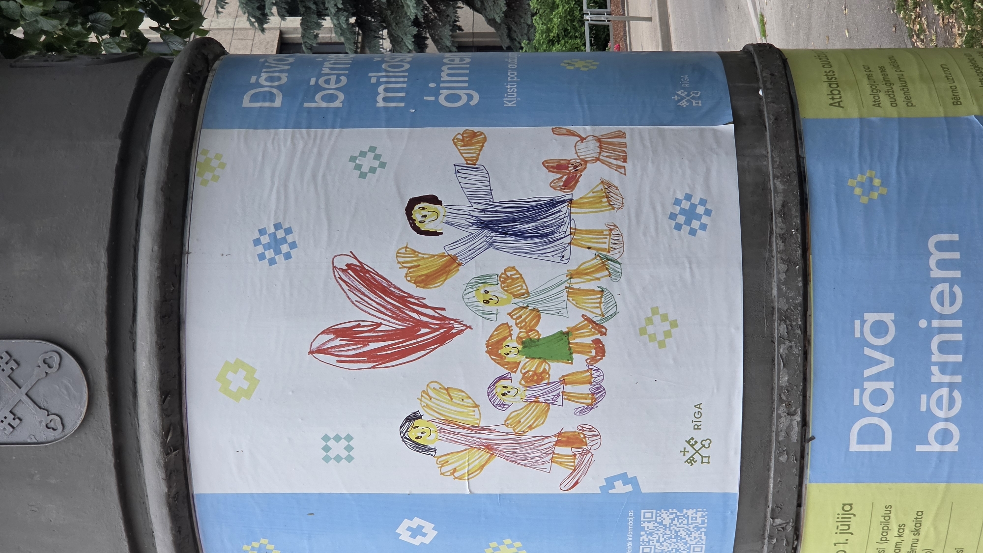 This is a photograph of a poster affixed to a cylindrical object, likely a public bin or pillar, in Riga, Latvia. The poster features a colorful, child-like drawing of a family with simplified figures, including two adults and three children, embracing or dancing. A large red heart is also prominently displayed. The background of the drawing is white, adorned with scattered, geometric patterns in various colors such as blue, yellow, and green.
The poster itself is split into sections with different colored backgrounds and text. The left section has a light blue background with the word "RĪGA" and a crest symbol. The central and right sections are also predominantly light blue, with white text and more geometric patterns. Some of the text is in Latvian, including "Dāvā bērniem" which translates to "Give to children" and "Atbalsts audžuģimenes" which means "Support for foster families". Other text provides information about allowances for foster families and mentions dates like "1. jūlija" (July 1st). A QR code is visible in the bottom right corner, suggesting a way to access more information. The overall scene appears to be outdoors, with glimpses of trees and a road in the background. The poster's cheerful illustration and supportive message suggest an awareness campaign or charity event. The image is captured in daylight, with no specific indication of the time of day or weather conditions.