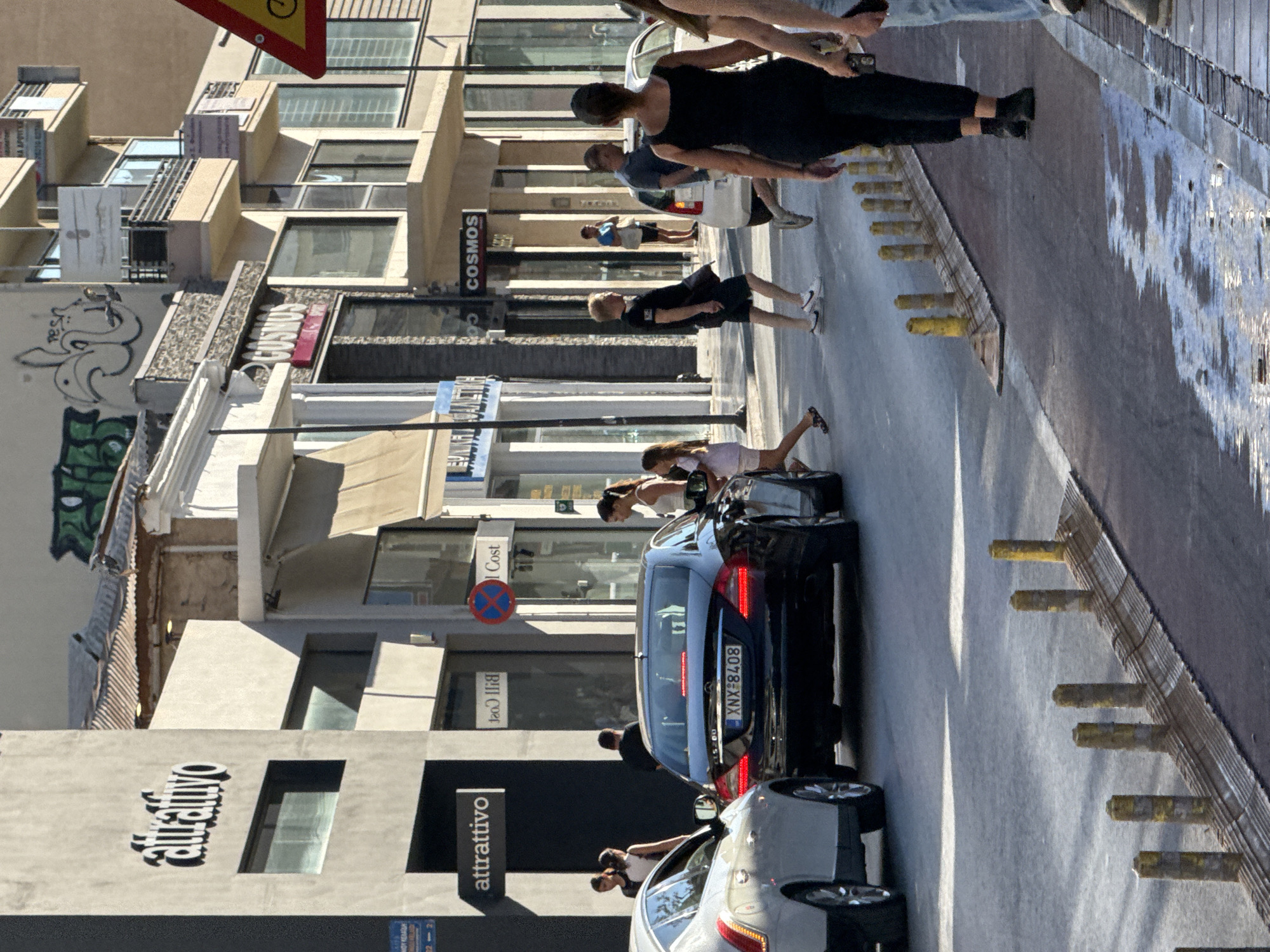 In Chania, Greece, on a sunlit street with buildings lining the right side, a black Mercedes-Benz sedan with license plate XNX 8408 is stopped in the roadway. Several pedestrians are crossing the street in front of the car and on the sidewalk. On the left side of the street, a white car is parked or moving slowly. Buildings have storefronts, including one with a sign reading "attraffivo" and another with "COSMOS." Some of the buildings display graffiti. The street is paved and appears wet in some areas, reflecting the sunlight. Bollards are visible along the edge of the sidewalk. The overall scene depicts a typical urban street with pedestrian and vehicle traffic under clear daylight conditions.