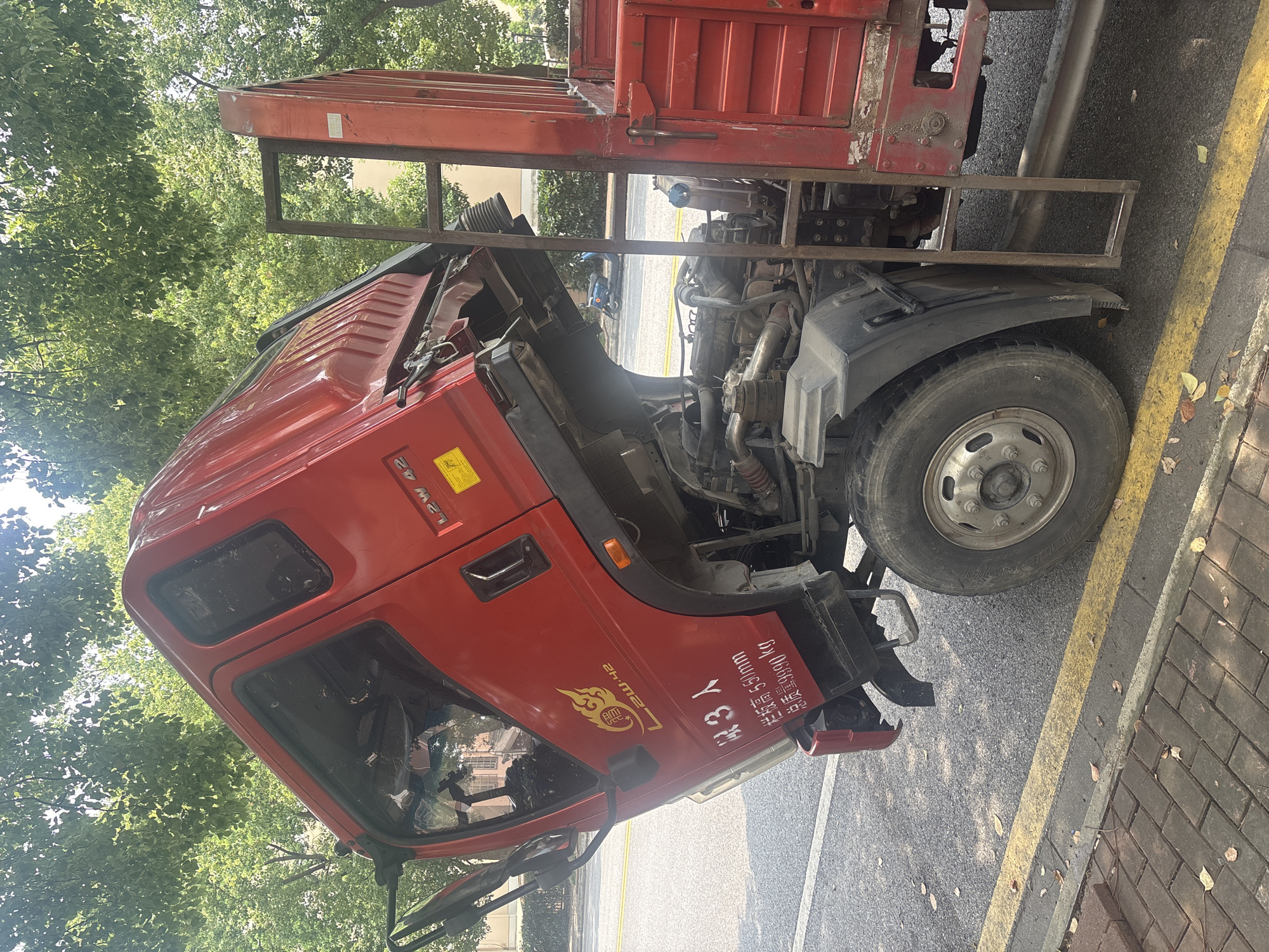 A red dump truck is parked on the side of a street in Suzhou Industrial Park, China. The truck's cab is tilted upwards, exposing the engine compartment. The truck is red and has a license plate that reads "L2W 42". There is a yellow sticker on the truck with Chinese characters. The truck is parked next to a sidewalk made of red bricks. There are trees in the background and a blue scooter is parked on the street behind the truck. The truck appears to be damaged, with some of the body panels dented and broken. The engine compartment is open, and some of the engine parts are visible. The truck has a ladder attached to the side of the bed.