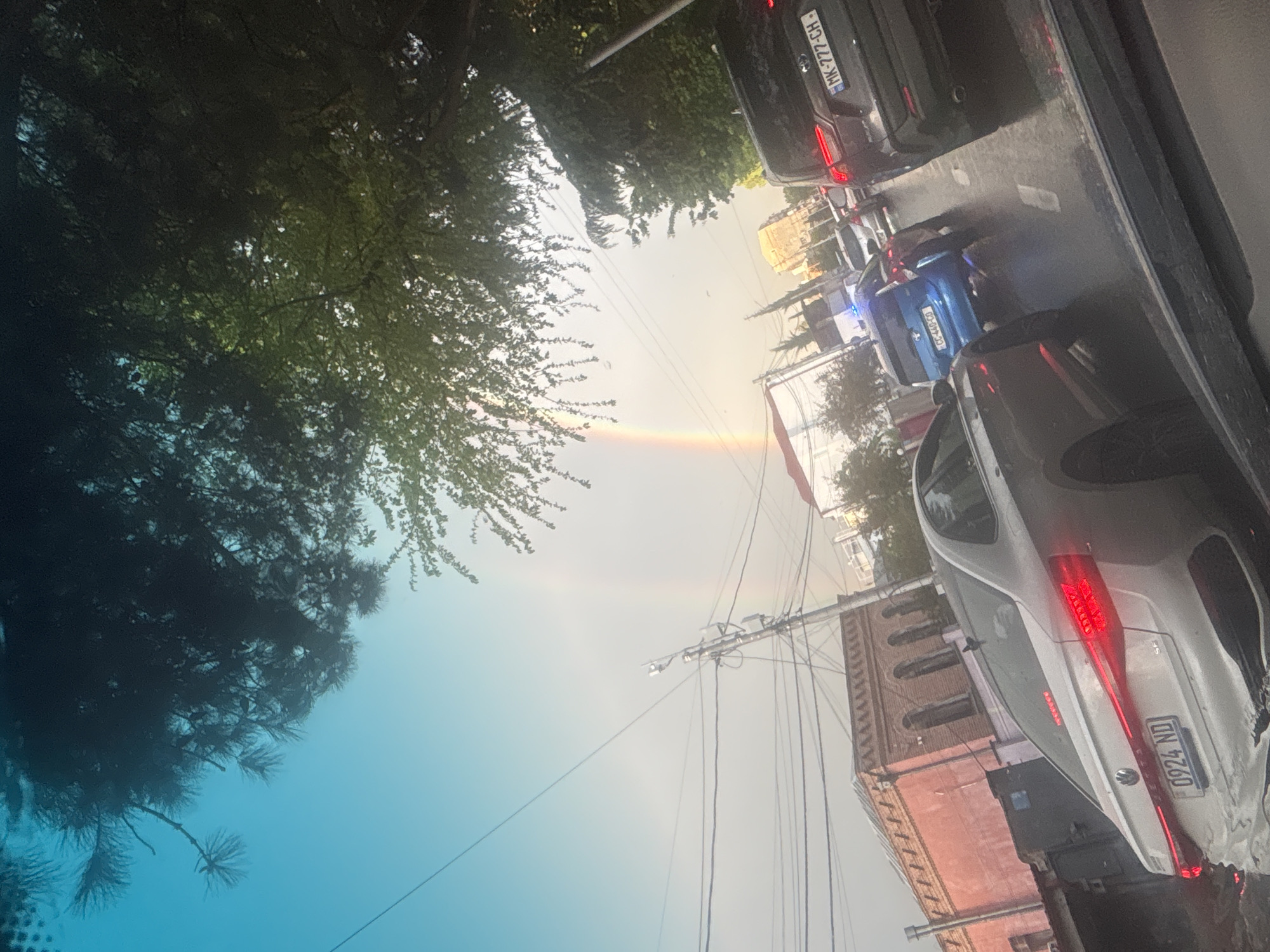 The image depicts a street scene in Tbilisi, Georgia, with a view of a partial rainbow arching across the sky. Traffic is present, including a silver car with the license plate "0924 ND," a blue car with the license plate "GG-697-VV," and a dark car with the license plate "HK-77-CH." A brick building with arched windows is visible on the right side of the frame. Overhead power lines stretch across the sky. A tree's foliage is seen on the left side, and the overall environment suggests an urban setting on a day with atmospheric conditions conducive to rainbow formation.