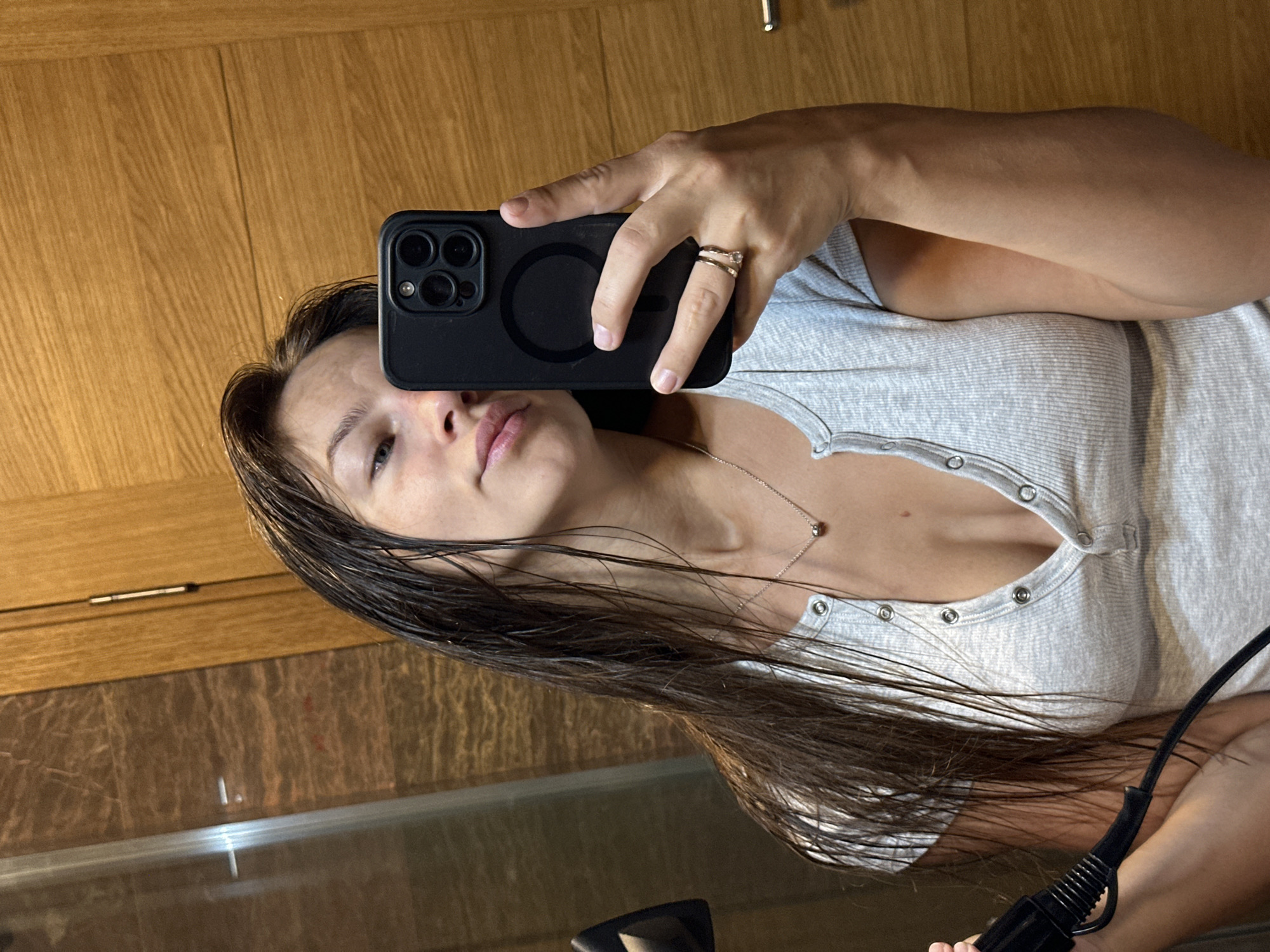A young woman with wet, dark brown hair is taking a selfie in what appears to be a bathroom. She is wearing a light grey, short-sleeved ribbed shirt with a buttoned neckline. She holds a black smartphone in her right hand, and a black hairdryer is visible in her left hand. A silver necklace with a small, spherical pendant is around her neck, and she is wearing two rings on her right ring finger. The background shows wooden paneling and tiled walls, suggesting an indoor setting. The overall scene is consistent with a person drying their hair after a shower. The specific location in Donji Pobori, Montenegro, is not visually discernible from the image itself.