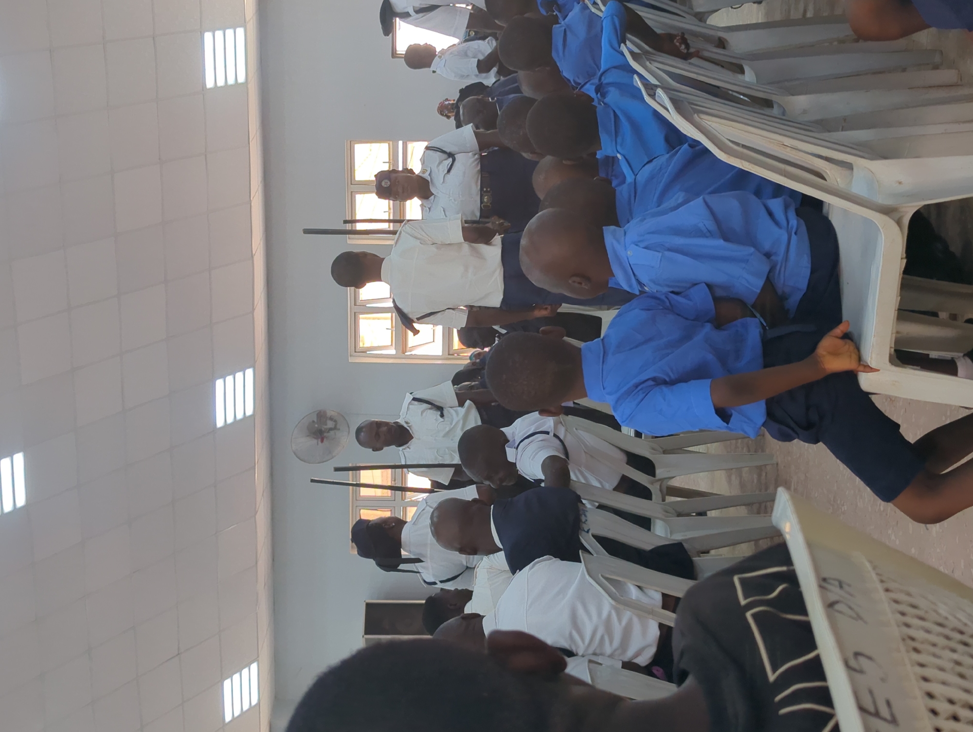 A room in Sabon Tasha, Nigeria, contains rows of plastic chairs occupied by people, some wearing white shirts and others in blue uniforms. Some stand while others sit. An electric fan is visible on the wall, and light enters through the windows. The tiled ceiling contains rows of fluorescent light fixtures.