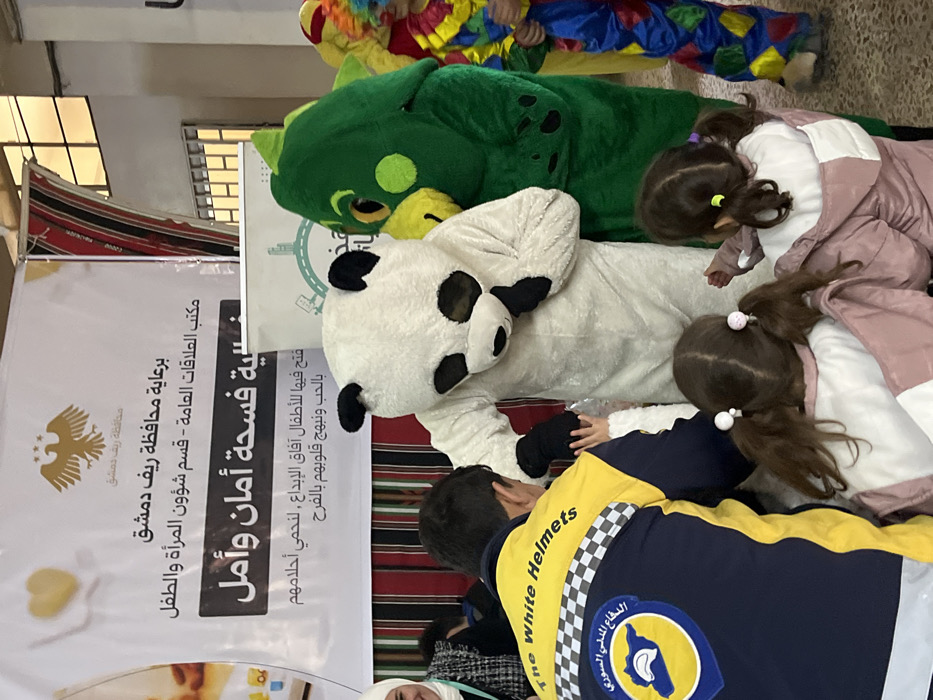 A group gathers around costumed mascots, including a panda and a green dragon. Two young girls in jackets are among them. A man wearing a White Helmets jacket is also visible. A banner in Arabic is displayed in the background, sponsored by the Rif Dimashq Governorate. The banner references opening horizons for children's creativity and protecting their dreams through love and joy.