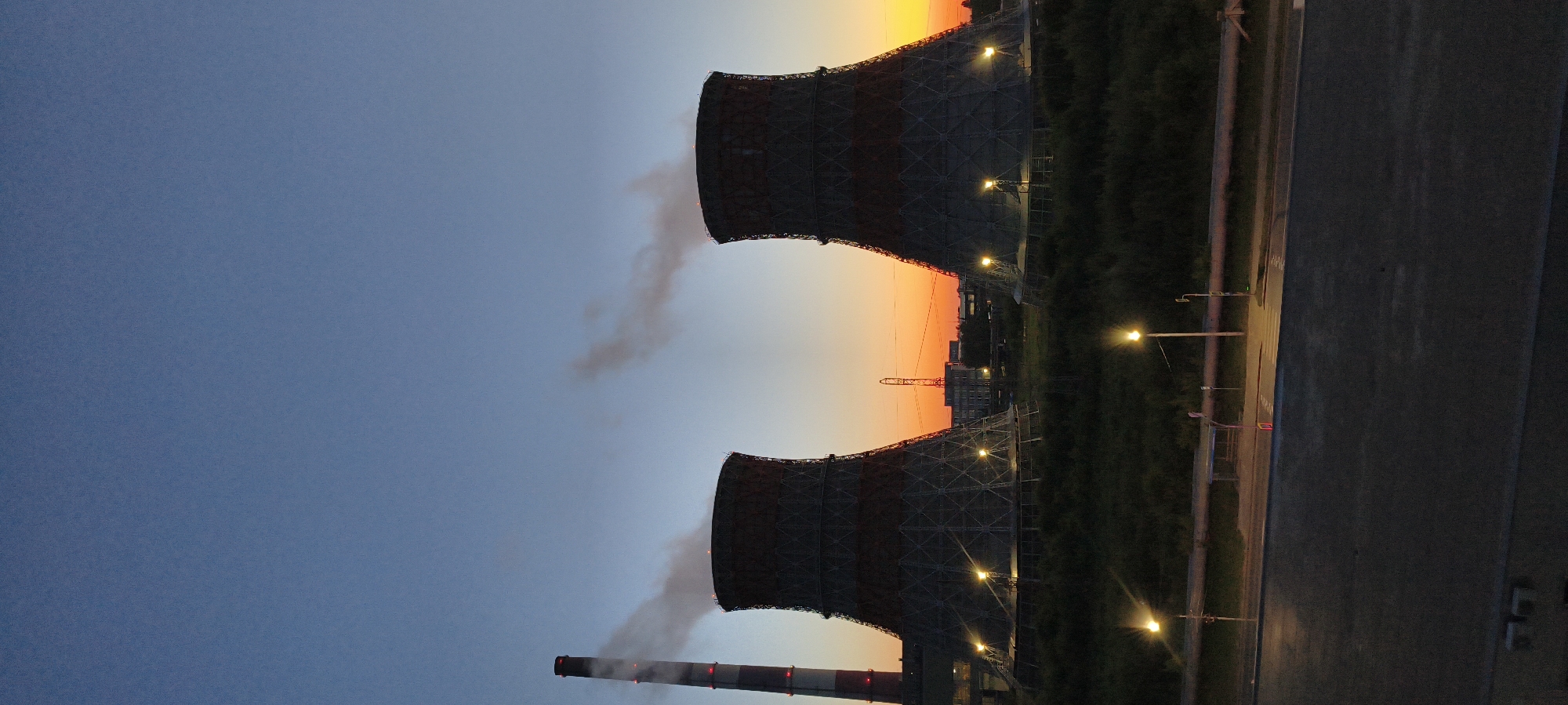 The image captures a twilight scene at a power plant, featuring two large, cylindrical cooling towers that dominate the center of the frame. The towers are tall and have a lattice-like structure supporting their wide bases and tapering tops. Red and grey sections are visible on the upper portions of the towers. Plumes of white steam or smoke rise from the tops of both cooling towers and from a red and white striped smokestack on the left side of the image. The sky above is a clear gradient of deep blue at the top, transitioning to a soft orange and yellow near the horizon, indicating either sunrise or sunset.
In the background, several low-rise buildings and a tall, slender tower structure with a lattice framework are visible against the colorful sky. The immediate area around the cooling towers is covered in dark green foliage, likely trees or bushes.
In the foreground, a paved area with streetlights illuminates a road and a crosswalk. The road appears to be asphalt and shows reflections from the streetlights. There are several lampposts with bright yellow lights positioned along the roadside. The overall atmosphere is one of industrial activity set against a natural twilight sky. The location is identified as Unknown, Unknown.