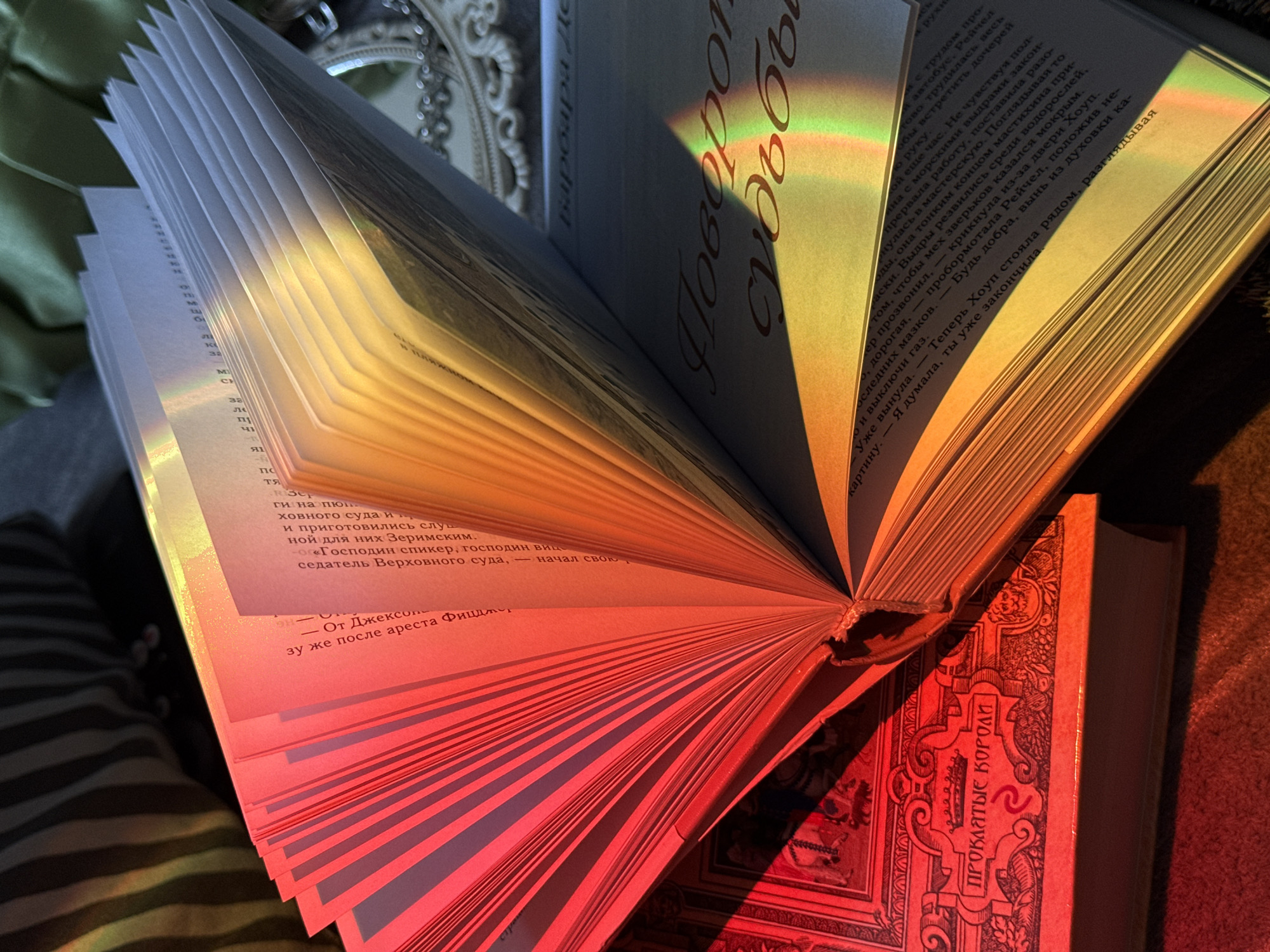 The image displays a close-up, high-angle view of two books in an indoor setting, illuminated by a striking rainbow light. The main subject is a large, open book whose pages are fanned out, creating a textured pattern. A vibrant spectrum of light, shifting from cool blue and green at the top to warm yellow, orange, and red at the bottom, falls across the pages, highlighting the text and the paper's texture.
On the right page of the open book, parts of a title in Cyrillic script are visible: "Поворо" and "судьбы," which likely form "Поворот судьбы" (Turn of Fate or Turning Point of Destiny). The left page contains more Russian text, including the name "Барбара Де" (Barbara de) and snippets discussing a judicial process, referring to "ховного суда" (Supreme Court), "господин спикер" (Mr. Speaker), and an event "после ареста Фицдти" (after Fitzdti's arrest). Another visible line mentions "Хоуп стояла рядом, разглядывая картину" (Hope stood nearby, looking at the picture).
Below the fanned book, another book, possibly a hardcover, is partially visible, its ornate reddish-orange cover catching the red portion of the rainbow light. On its cover, the Cyrillic title "ПРОКЛЯТЫЕ КОРОЛИ" (THE ACCURSED KINGS) is clearly discernible within an elaborate decorative frame.
In the blurred background, an ornate, possibly silver-colored decorative frame or mirror is partially visible, alongside a hint of green fabric. The intense, refracted light creates a magical and cozy atmosphere, suggesting a quiet moment of reading or contemplation during daytime. No people are visible.