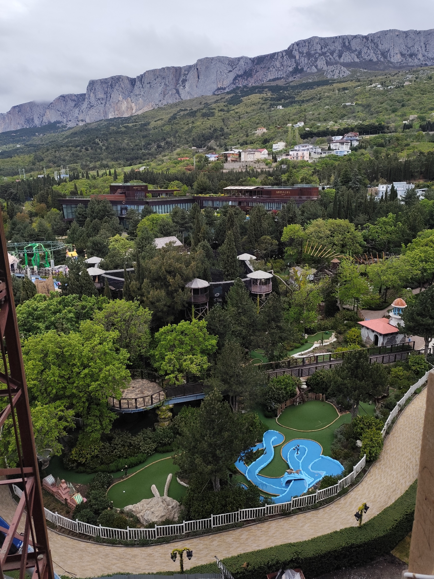 The image captures a high-angle view of a landscape in Симеизский поселковый совет, Ukraine. Dominating the background is a large, rugged mountain range under a cloudy sky. In the midground, there is a dense mix of trees interspersed with buildings, including a large modern structure with a dark roof. In the foreground, a mini-golf course is visible, featuring a winding blue water feature, artificial green grass, and a white picket fence. A paved walkway runs alongside the mini-golf course. Some construction or maintenance equipment is present in the lower left corner of the image. No people are clearly visible.