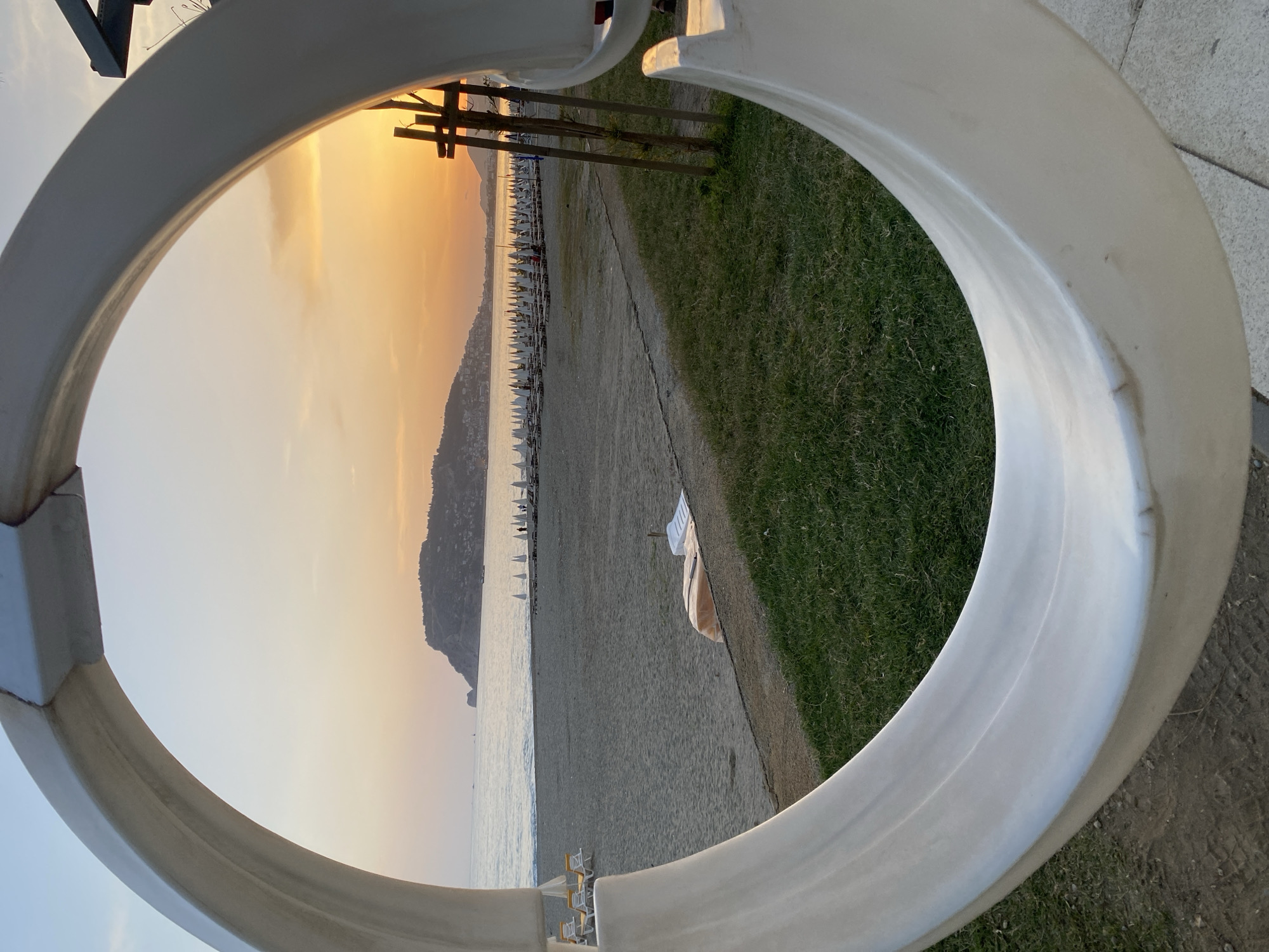 The image shows a beach scene framed by a large, white, circular structure. The beach is lined with numerous white umbrellas and lounge chairs. A small, mountainous landmass is visible in the distance. The sky is bright with the colors of a sunrise or sunset. A patch of green grass is visible along the edge of the beach. The structure appears to be part of a public art installation or architectural feature. The location is Alanya, Turkey.