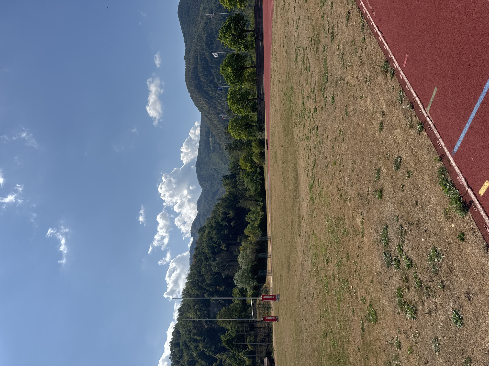 The image depicts an outdoor sports field, likely a rugby or soccer field, with a running track in the foreground. The field itself is predominantly covered in dry, brown grass, with patches of green weeds. In the background, a mountain range covered in dense green trees forms a natural backdrop. Several flags are visible on poles in the middle ground, and a set of rugby goalposts with red padding is located on the left side of the field. The sky is a clear blue with scattered white clouds. The scene appears to be located in Saint-Claude, France, given the context. The overall environment suggests a sunny day in a mountainous region.
