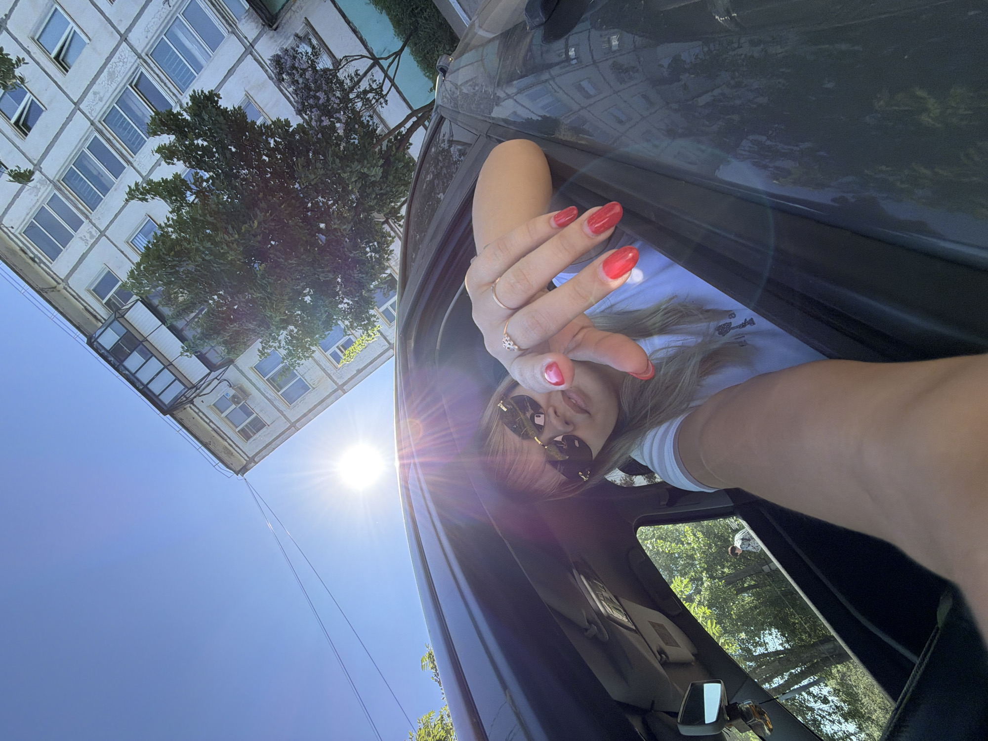 A woman with sunglasses and red nail polish on her fingers is leaning out of the open sunroof of a dark car. The image is taken from a low angle, looking up at the woman. Outside the car, there is a blue sky and a building with many windows and a tree in front of it. Based on the architecture of the building and some vegetation, the location is likely Chaikovsky, Russia.