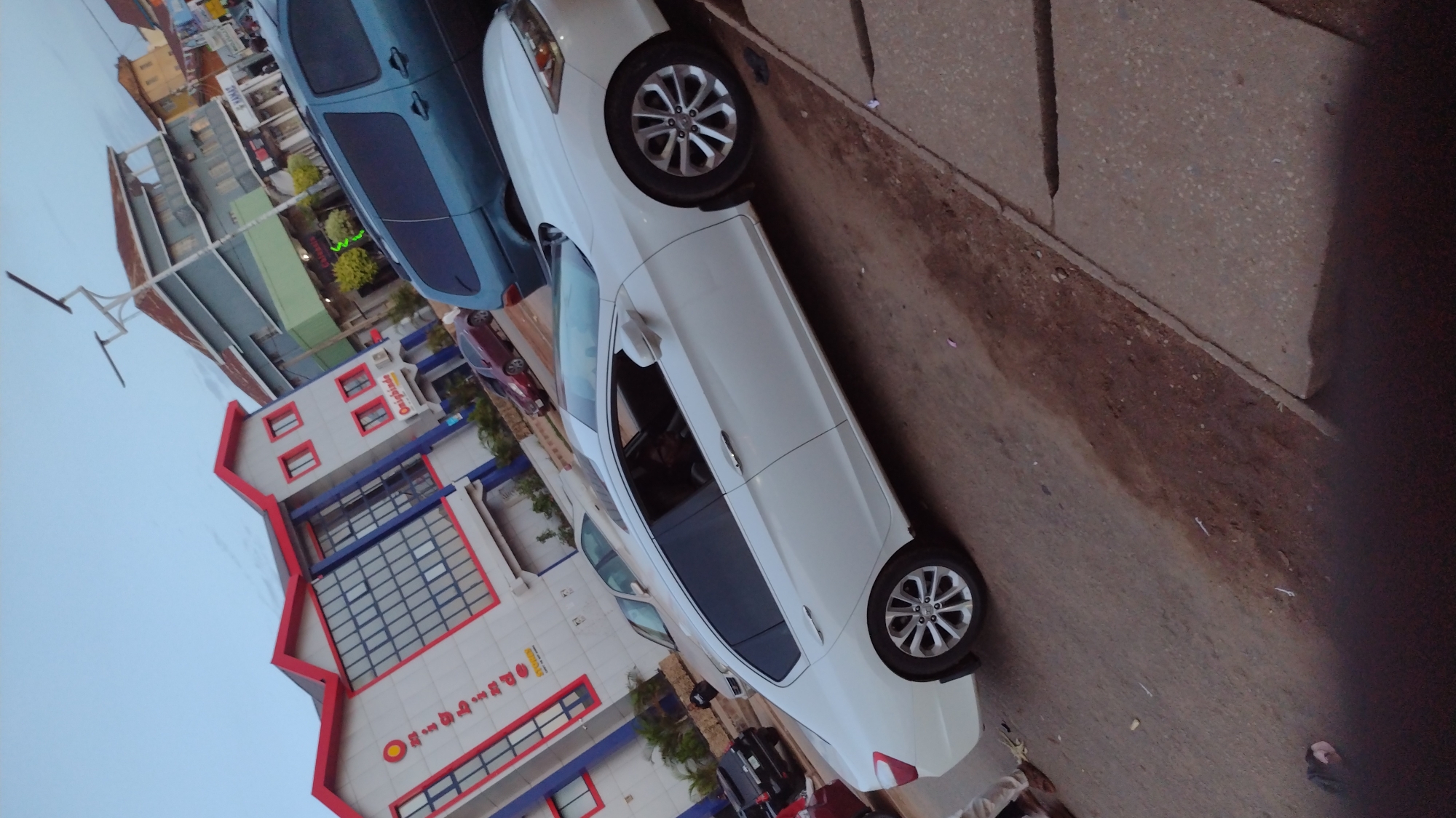 This image, rotated 90 degrees counter-clockwise from its natural orientation, captures a daytime street scene in Jos, Nigeria.
In the foreground, a white sedan, resembling a Hyundai Elantra, is prominently parked on the side of the road. It features distinctive alloy wheels. Inside, a person is faintly visible in the driver's seat. Behind the white sedan, a blue van or minivan is parked, and further down the street, a reddish-brown sedan is also visible. A black motorcycle or scooter is partially obscured behind the white car's rear wheel.
On the left side of the scene, a multi-story building with a striking red and light grey/white facade stands out. The upper section of the building displays the text "Omoigboade" in white letters on a red background. On the lower part of the facade, a larger sign clearly reads "Omoigboade GROUP OF SCHOOLS" in yellow text, accompanied by a circular logo featuring "OS" and the words "OMO SCHOOLS". Further back, another building has a sign with faint green lettering, possibly "CANNON". A utility pole or street light structure is visible on the far left.
The sky appears overcast or hazy, suggesting it is daytime, possibly late afternoon, given the soft lighting. The road surface consists of asphalt, bordered by a dirt and gravel shoulder adjacent to a concrete sidewalk. A person's foot is partially visible in the bottom right corner of the frame, indicating someone is standing or walking nearby. The scene depicts a typical urban street environment with parked vehicles and commercial or educational buildings.