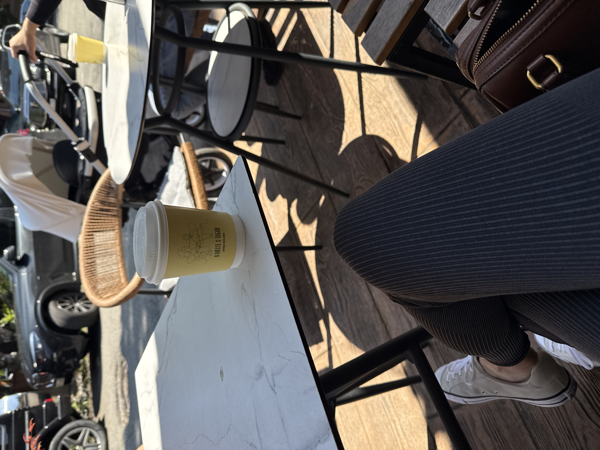The image depicts a scene in Sochi, Russia, featuring a person seated at an outdoor café. A partially visible brown leather purse rests on the table next to the person, who is wearing dark gray striped pants and light gray sneakers. A yellow paper cup with Russian text "ВМЕСТЕ С ТОБОЙ" ("Together with you") and a drawing of a cook holding a tray is on the table. Other tables and chairs are visible, as is a stroller and a car parked nearby. Sunlight casts shadows on the wooden floor of the cafe.