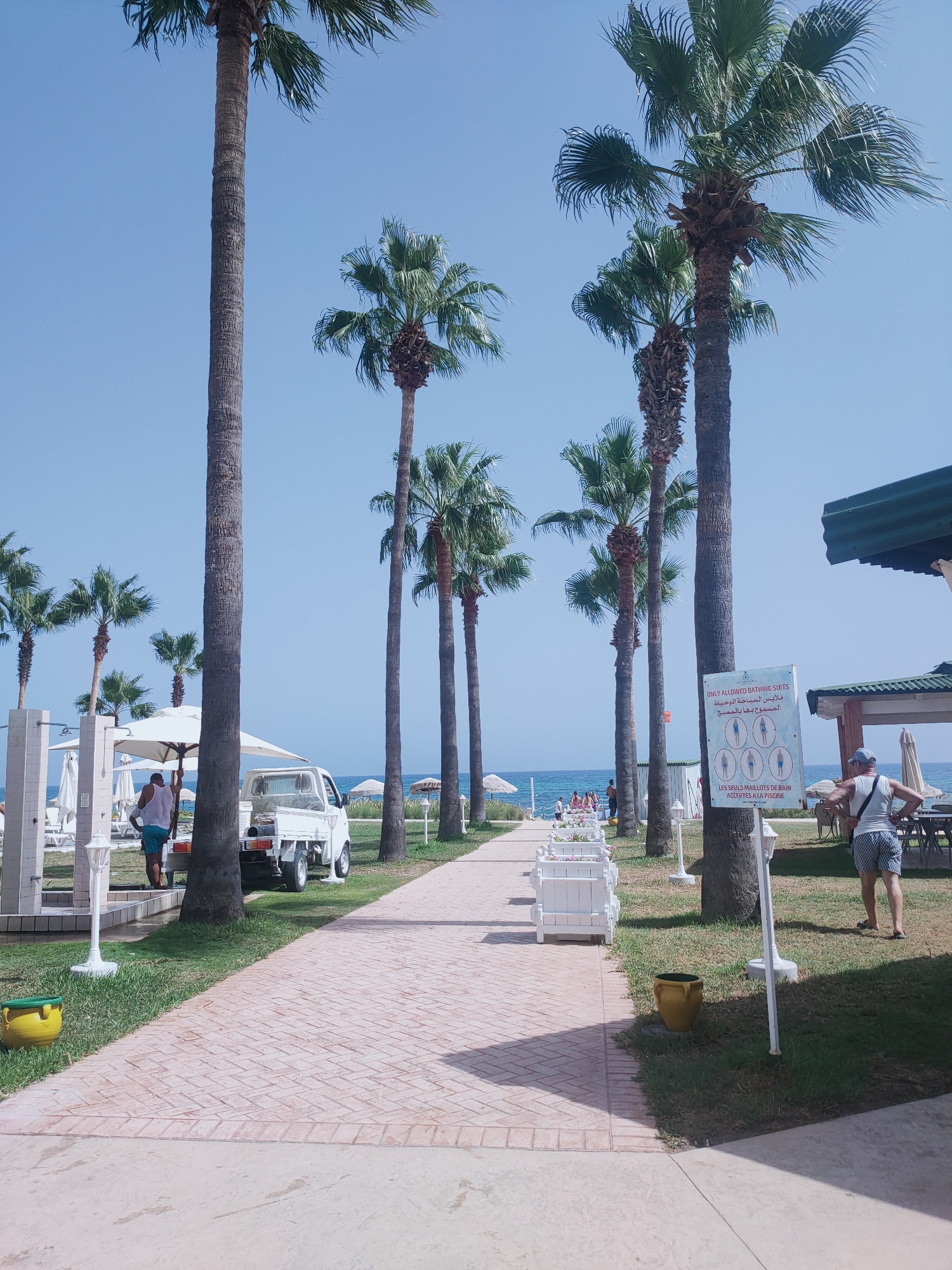 The image captures a sunny day in Nabeul, Tunisia, featuring a paved walkway leading towards a beach. Tall palm trees line the path, with a clear blue sky overhead. On the left side of the walkway, there's an outdoor shower area with white structures. A white truck is parked near the walkway, and a man in a white tank top and patterned shorts stands near it, appearing to be attending to something. Further down the path, beach umbrellas and lounge chairs are visible, suggesting a resort or public beach area. In the background, the blue sea stretches out to the horizon, with a few people visible on the beach. A signpost near a palm tree displays rules in both English and Arabic, indicating that only bathing suits are permitted. The scene is tranquil and depicts a typical beach resort atmosphere.