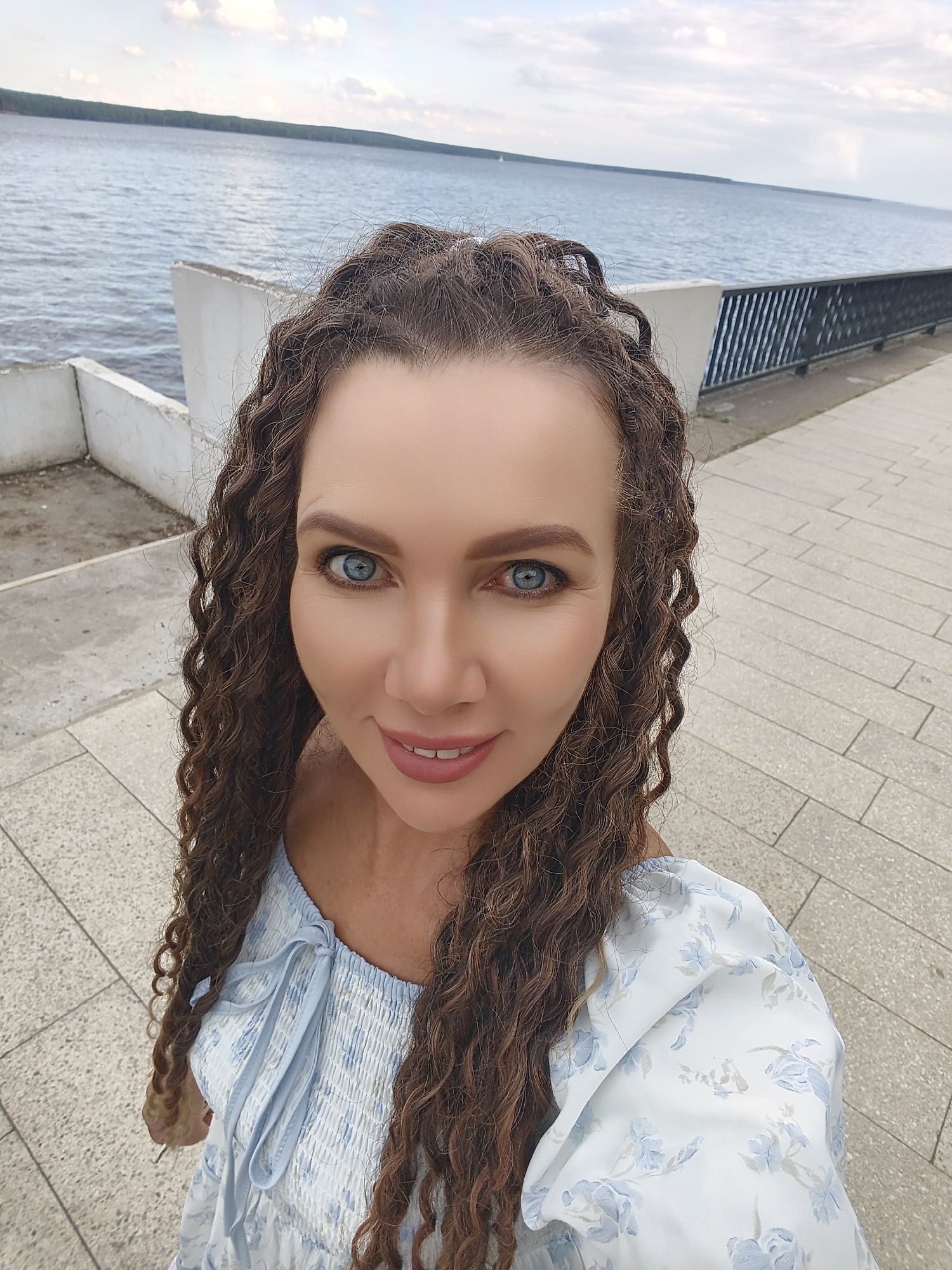 This is a selfie of a woman with long, curly brown hair and bright blue eyes, taken outdoors on a cloudy day. She is smiling, with her mouth slightly open, and wearing a light blue and white floral print dress with smocking on the bodice. The background shows a paved walkway, a body of water with a distant tree line, and a white concrete structure. A black metal railing runs along the edge of the walkway. The overall impression is of a casual, pleasant outing by the water. The time of day appears to be afternoon or early evening, given the lighting. The location context is Chaikovsky, Russia. There is no visible text in the image.