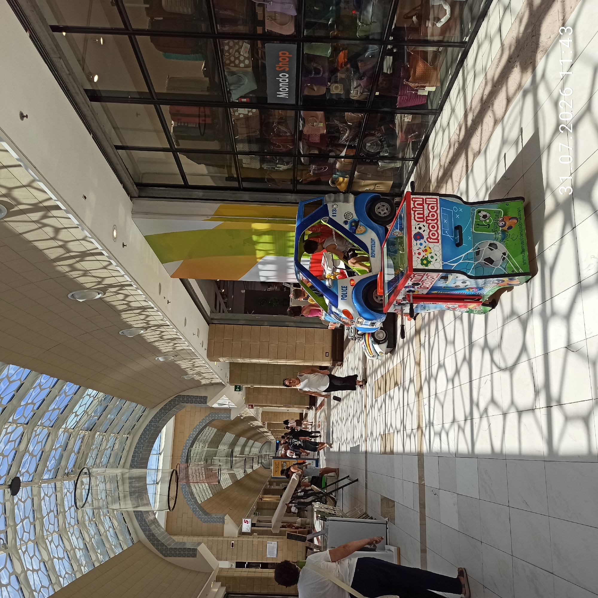 The image depicts a view inside a modern shopping mall in Modugno, Italy. The architecture features a high, arched ceiling with a transparent, patterned roof that allows natural light to filter in, casting hexagonal shadows on the white tiled floor. The mall's interior is lined with shops, one of which, "Mondo Shop," is visible with its display of handbags.
In the foreground, a woman with dark hair, wearing a white t-shirt and dark pants, walks away from the camera. To her right, a colorful, child-sized arcade game, a "mini football" table, stands on the tiled floor. Adjacent to it is a blue and white toy police car with children seated inside.
Further down the mall's corridor, other shoppers are seen walking, including a woman in a white top and dark pants approaching the camera, and several people in the distance. The overall impression is of a bright, airy, and active shopping environment. A timestamp at the bottom right corner reads "31.07.2026 11:43".