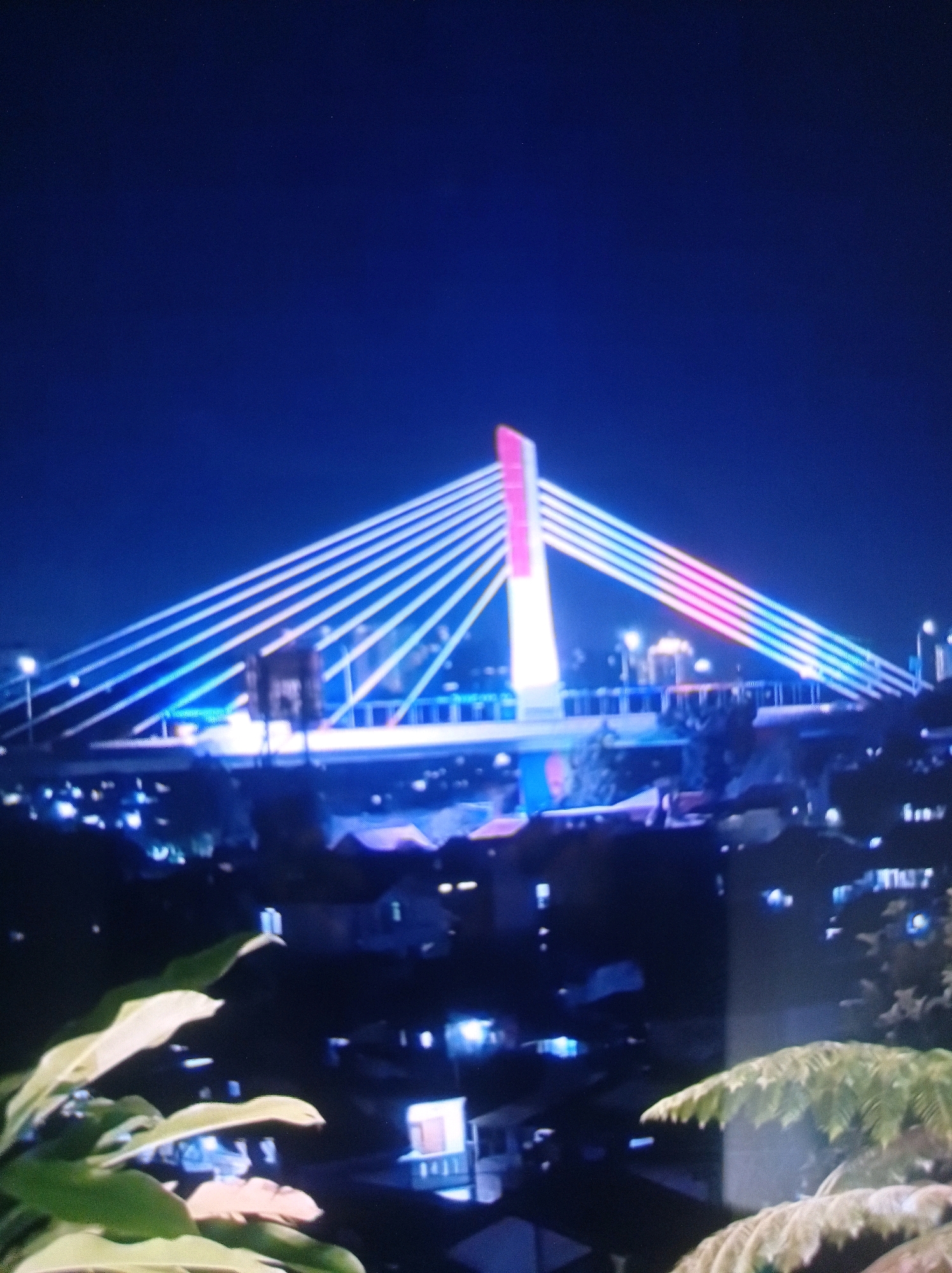 The image captures a nighttime view of the Pasupati Bridge in Bandung City, Indonesia. The bridge is illuminated with a vibrant display of lights, featuring a prominent pink and white pylon and cables that glow with blue and pink hues. In the foreground, out-of-focus foliage suggests a vantage point from a higher elevation, likely a hillside overlooking the city. The background reveals the soft, scattered lights of houses and buildings, indicating a residential or urban area spread across the landscape beneath the illuminated bridge. The overall scene conveys a festive and picturesque urban nightscape.