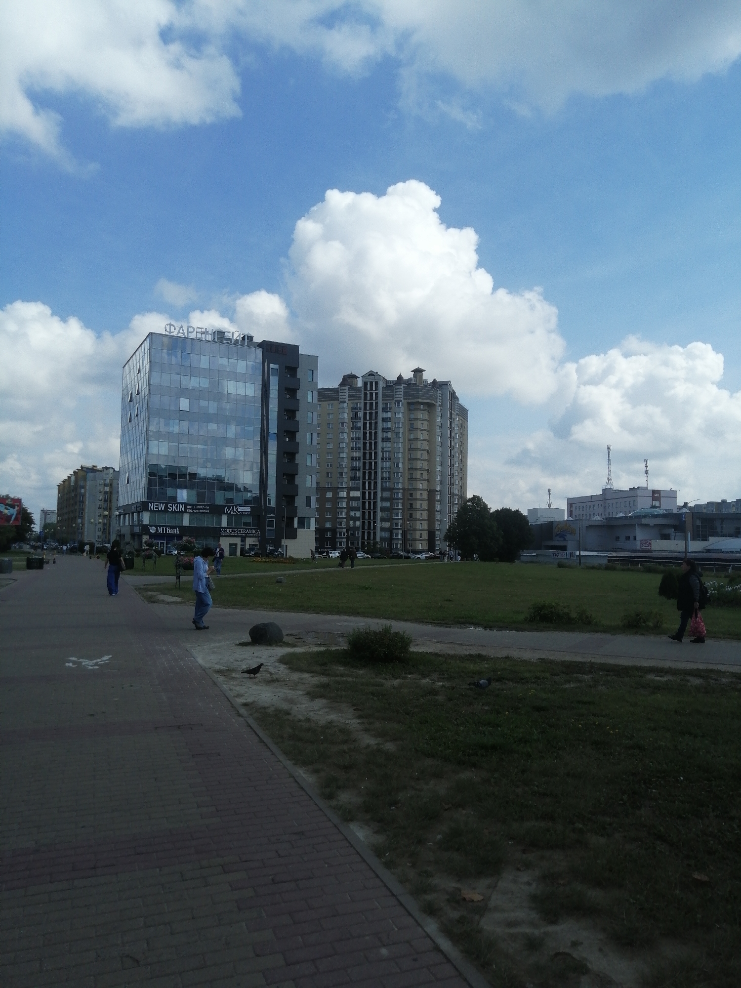This is a street-level, eye-level, full-shot photograph taken in Minsk, Belarus, on a cloudy but bright day. The foreground is dominated by a paved walkway made of reddish-brown bricks. A few pigeons are visible on the walkway and on the adjacent grass. To the left of the walkway is a grassy area.
In the mid-ground, a modern glass-fronted building with the word "ФАРМЦІЯ" (Farmatsiya) in large silver letters across its top facade is prominent. Below this, signs indicate "NEW SKIN," "MK," "MTBank," and "MODUS CERAMICA." To the right of this building is a taller, multi-story residential building with a curved, decorative facade and numerous balconies. Further in the distance, other buildings of varying heights and architectural styles can be seen, including a long, low-rise commercial or industrial building with antennas on its roof.
Several people are visible walking along the paved walkway. A person in a blue shirt and jeans is walking towards the right. Further left, a person in a dark top and blue pants is walking away from the camera. Another individual in dark clothing, carrying a red bag, is walking towards the camera on the far right. The sky is filled with large, fluffy white clouds against a bright blue background. The overall impression is of a modern urban environment with a mix of commercial and residential structures.