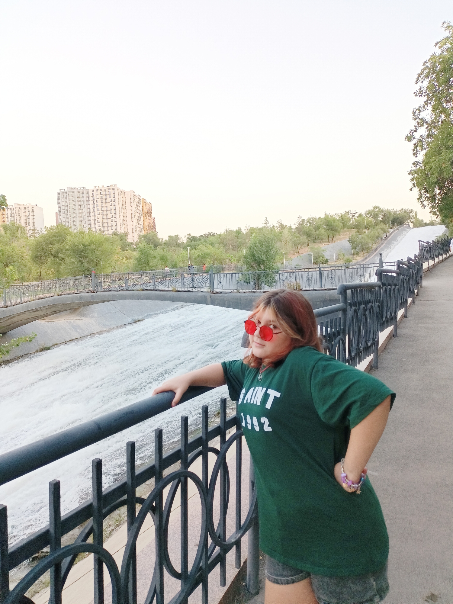 A young female with reddish-blonde hair and red-rimmed sunglasses leans against a black metal railing. She is wearing a dark green t-shirt with white lettering that appears to read "SAINT 1992" and dark denim shorts. A colorful bracelet is visible on her left wrist. The background shows a concrete canal with rushing water, a pedestrian walkway with a black metal fence, and a bridge in the distance. Beyond the canal and bridge, there are trees and several tall apartment buildings, characteristic of the city of Almaty, Kazakhstan. The sky is overcast with a light, hazy glow. The overall scene appears to be a public park or recreational area.