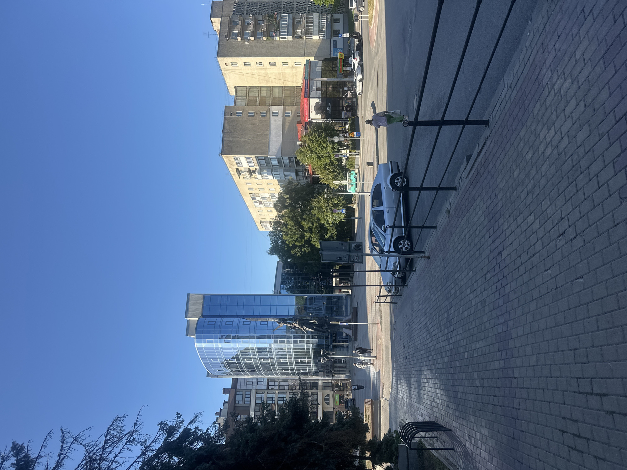 The image shows a street scene in Ternopil, Ukraine, on a clear, sunny day. In the foreground, a paved walkway made of grey interlocking bricks runs alongside a black metal fence. To the left of the walkway is a bicycle rack. Beyond the fence, a row of parked cars is visible, including a silver sedan. Further down the street, a woman in a light-colored top and green pants walks away from the camera, carrying a bag.
Dominating the mid-ground is a modern, multi-story building with a curved glass facade, reflecting the blue sky. A statue is prominently displayed in front of this building. To the left of the glass building, older, multi-story apartment buildings are visible. Across the street, more apartment buildings, some with balconies, line the horizon. A bus stop with a turquoise minibus is also visible in the distance. The street is paved, and trees are scattered throughout the scene, some with green foliage. The overall impression is an urban environment bathed in sunlight.