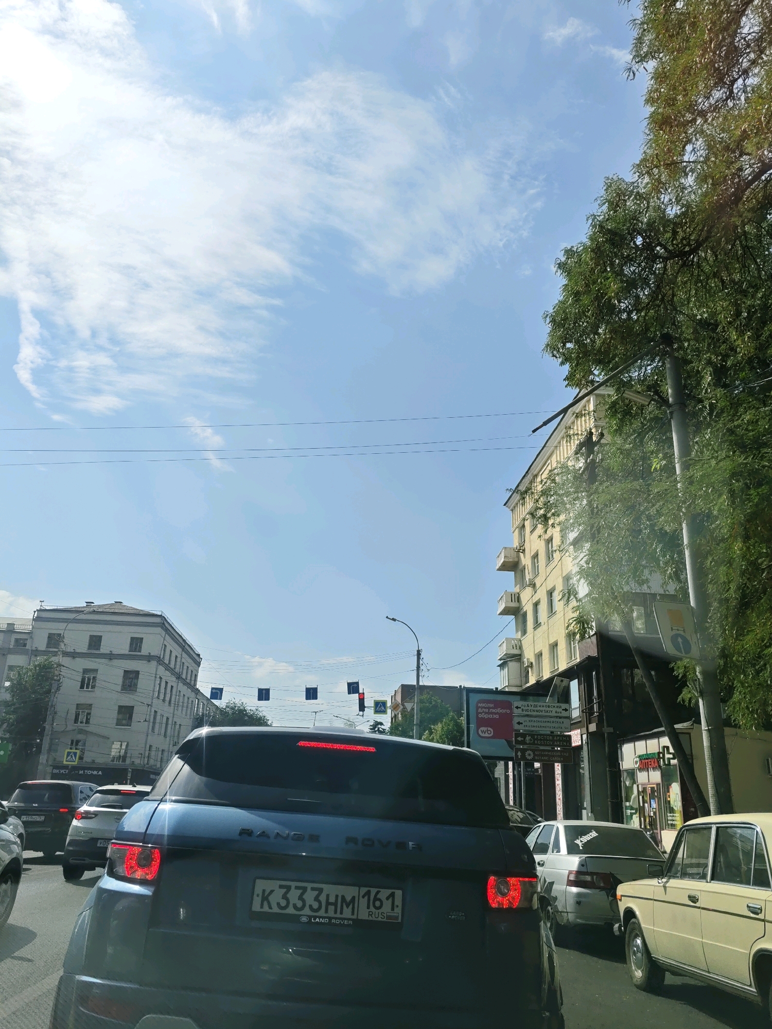 The image captures a traffic jam on a sunny day in Rostov-on-Don, Russia. In the foreground, a dark blue Range Rover is stopped, its brake lights illuminated. The license plate reads "K333HM 161 RUS," indicating it is registered in the Rostov region. Several other cars are visible in the queue, including a white sedan with a sticker on its rear window and a light-colored vintage car.
The background features apartment buildings and trees lining the street. Overhead wires crisscross the sky, which is partly cloudy with a bright sun. Signs with Russian text are visible on the side of the road, including directional signs pointing to "BUDENNOVSKY AVE" and "ROSTOV ARENA." The overall scene depicts a typical urban environment experiencing a traffic standstill.