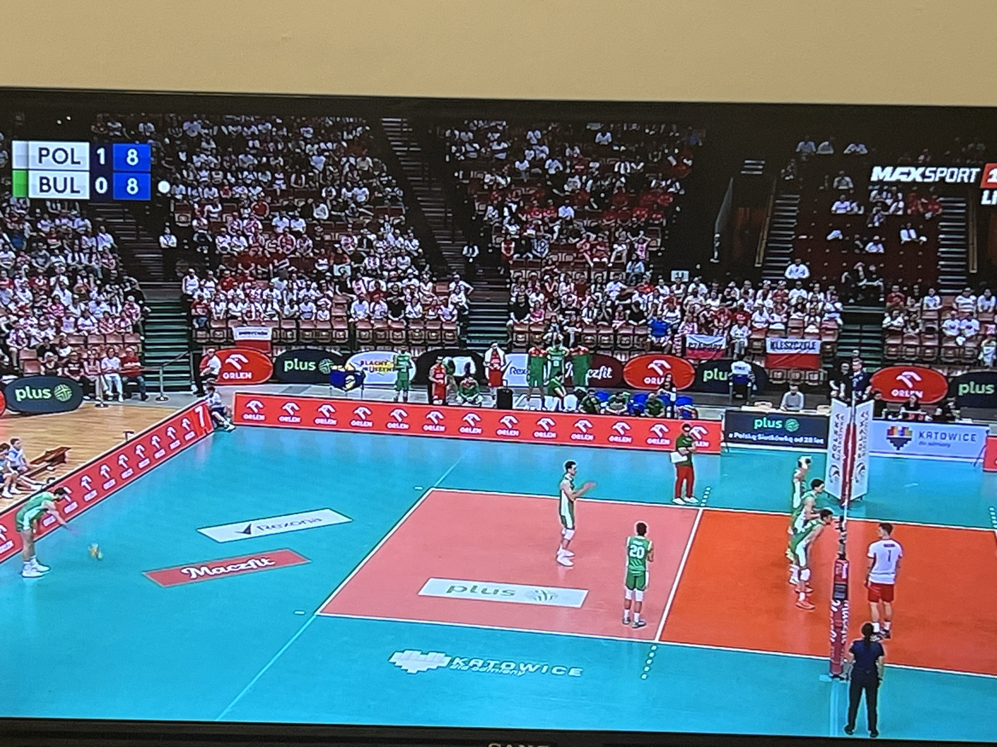 The image shows a volleyball match between Poland (POL) and Bulgaria (BUL). The score is 18-8 in favor of Poland. The match is taking place in Katowice, Poland. There is a large crowd in attendance. Players from both teams are on the court. The court is marked with advertisements from companies such as Plus, Orlen, Rexona, and Maczfit. A referee stands near the net. No information can be determined about Gabrovo, Bulgaria from this image.