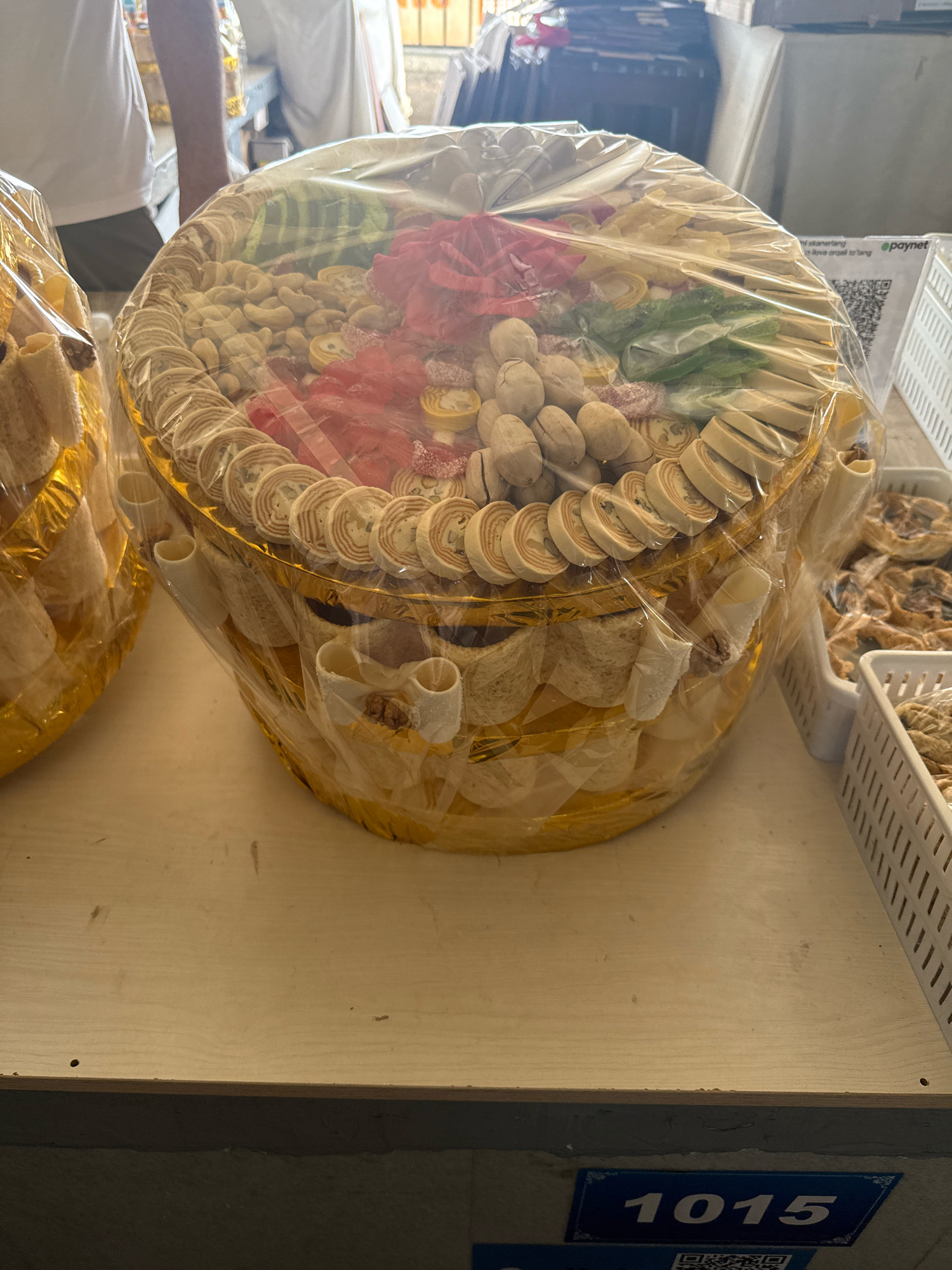 The image shows a large, round platter filled with assorted food items, possibly dried fruits, nuts, and candies, covered in clear plastic wrap. The platter itself is gold-colored and sits on a wooden surface. To the left, another similar platter is partially visible. In the background, there are glimpses of people and a store environment, including white baskets containing baked goods. A QR code and the word "epaynet" are visible on a sign in the background, suggesting a commercial setting. The scene appears to be inside a shop or market stall. The visible numerical sign "1015" may indicate a stall number or product identifier. The general context suggests a food vendor in a market in Tashkent, Uzbekistan.