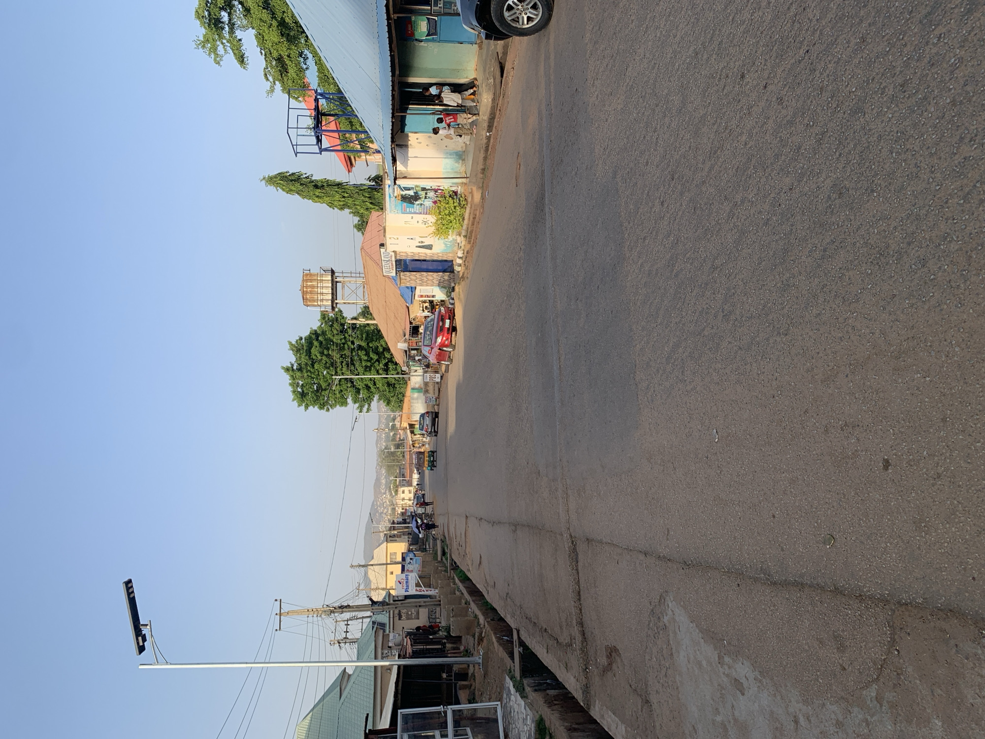 The image captures a street view in Kabong, Nigeria. The road, made of asphalt, stretches into the distance. Buildings line one side of the street, showing diverse architectural styles, including corrugated metal roofs and painted facades. Some shops have awnings. Various vehicles, including cars and motorcycles, are on the road. In the distance, the scene ends with hills and houses. The sky is clear. Utility poles are visible along the street. Several individuals are present along the street.
