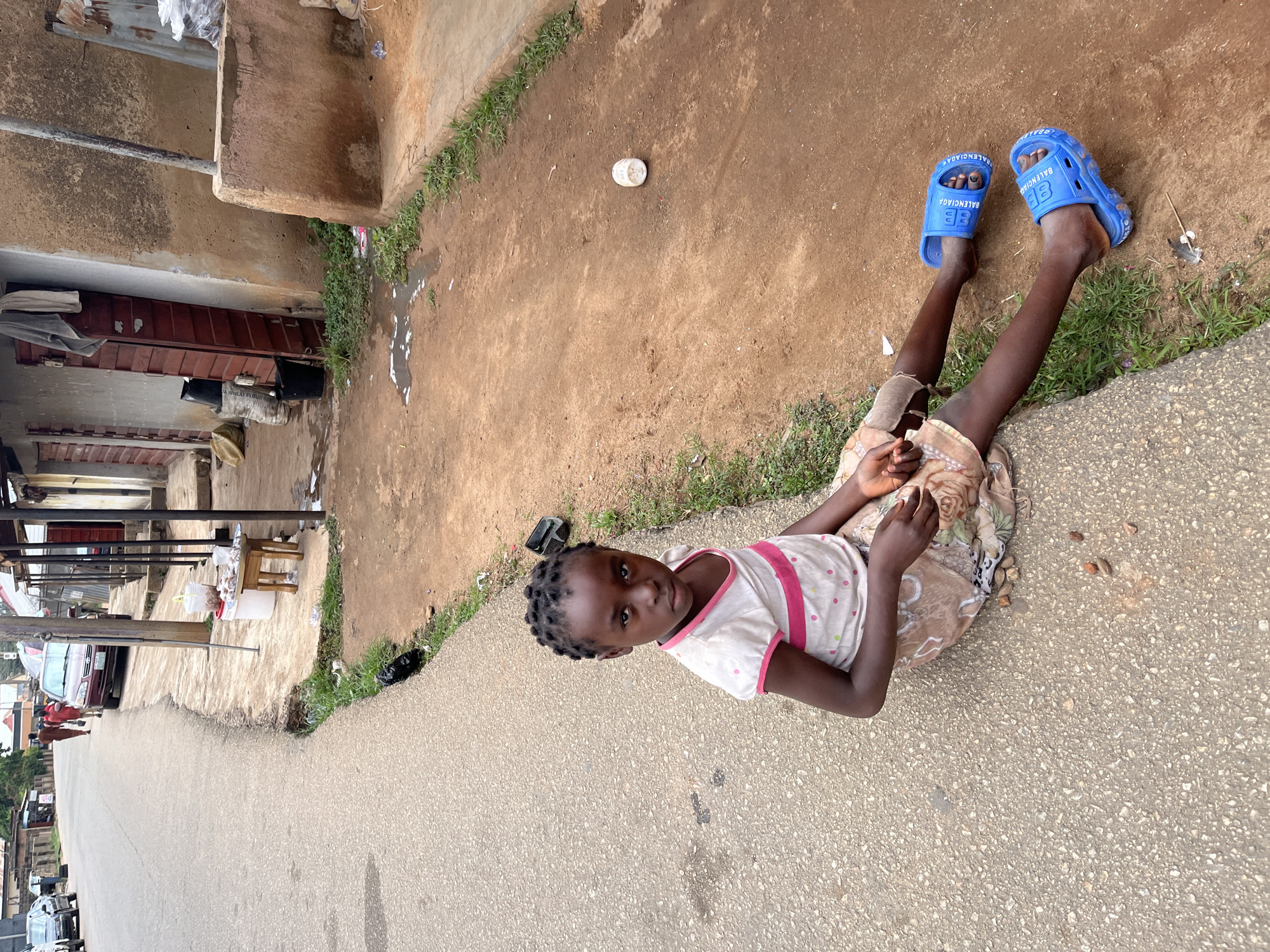 A young girl is positioned at the intersection of a paved road and a dirt shoulder in Jos, Nigeria. She is seated directly on the gray asphalt, with her legs extending onto the reddish-brown earth. The girl, estimated to be between six and ten years old, has dark skin and her hair is styled in tight, small braids. She wears a white t-shirt with pink trim and two horizontal pink stripes, adorned with small multi-colored polka dots, and a light brown patterned skirt or wrap. Her feet are clad in bright blue "Balenciaga" branded slide sandals. Her gaze is directed towards the viewer with a neutral expression.
To her right, on the dirt, a few small, reddish-brown stones or nuts are scattered. Further right, a rudimentary street stall is visible, consisting of a small wooden stool supporting a metal tray with white, round food items (possibly peeled tubers) and two white plastic buckets. Behind this setup, simple, unpainted or light brown rendered buildings with wooden doors line the dirt path. Laundry is hanging near one doorway. Patches of green grass and weeds grow along the edge where the pavement meets the dirt, interspersed with general litter like plastic bags and puddles.
The paved road, appearing to be a two-lane street, recedes into the background, curving slightly to the left. A red sedan and another vehicle are parked further down on the right side. Two individuals are seen walking away from the camera on the paved road in the distance. The overall scene depicts a functional urban street environment.