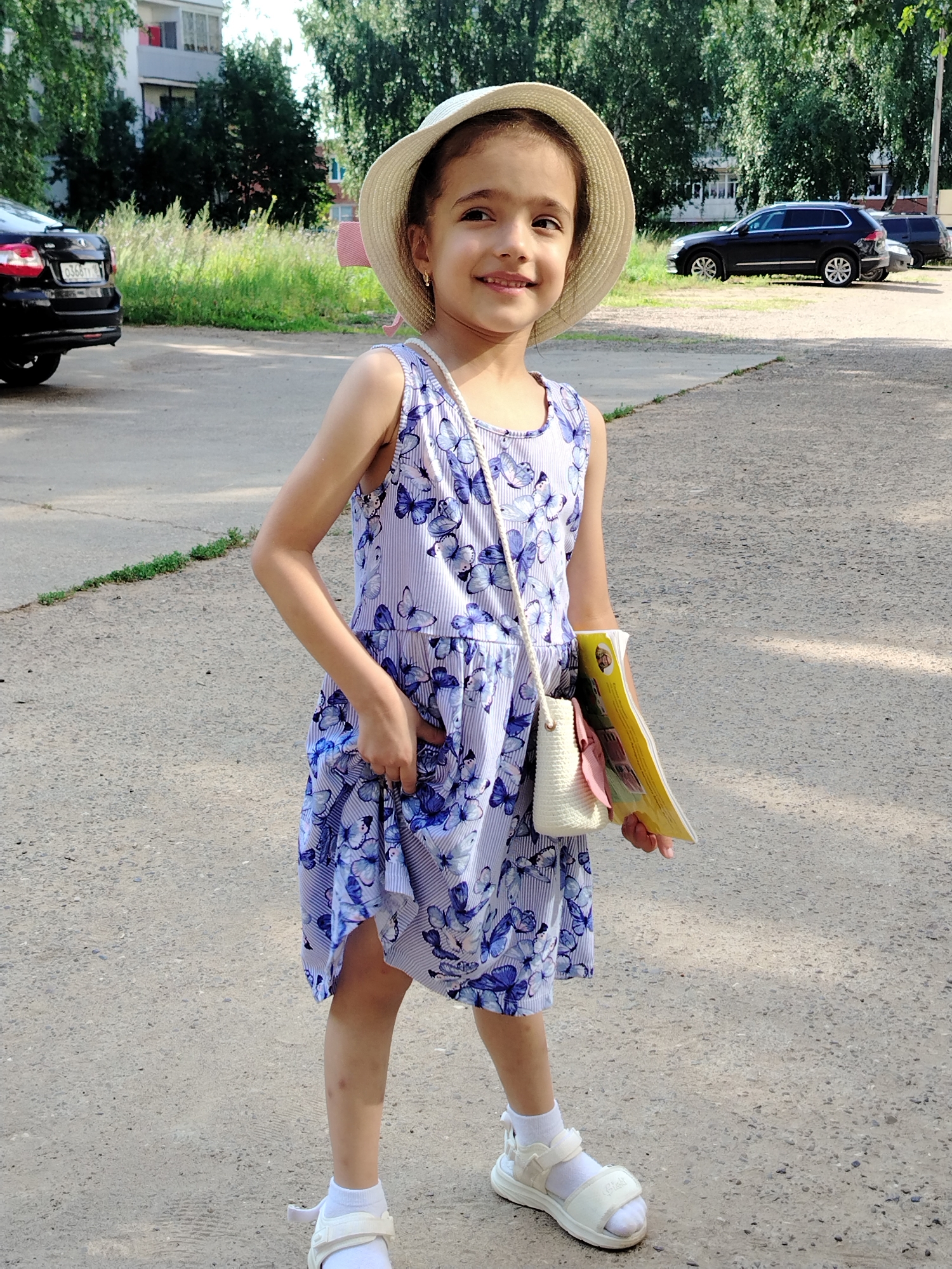 In Noviy, Russia, a young girl stands outdoors on a paved surface, likely a street or parking lot. She is wearing a light-colored straw hat, a sleeveless blue and white striped dress adorned with a butterfly pattern, and white sandals with white socks. The girl is holding a yellow book and a small white purse with a pink ribbon. Her right hand is in her dress pocket. Behind her, several cars are parked, including a black SUV. The background features trees and a residential building. The lighting suggests it is daytime.