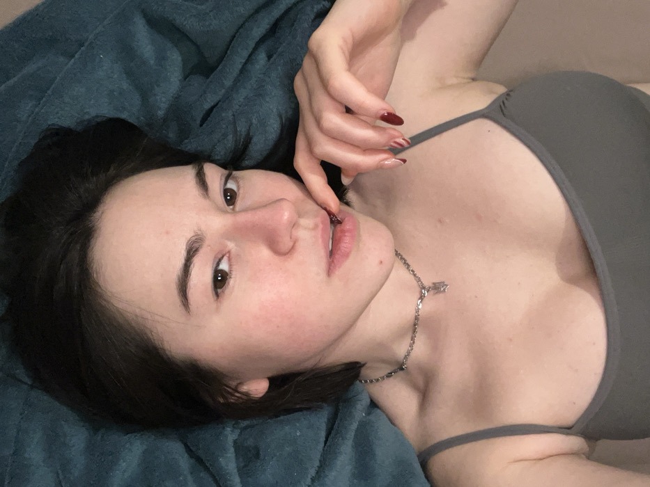 A woman with dark hair is lying on a blue blanket. She is looking at the camera with a playful expression and has her fingers on her lips. She is wearing a gray tank top, and a silver chain necklace with a small pendant. Her nails are painted with a dark red color, and some have a light pattern on them. The lighting is soft and even, highlighting her features.