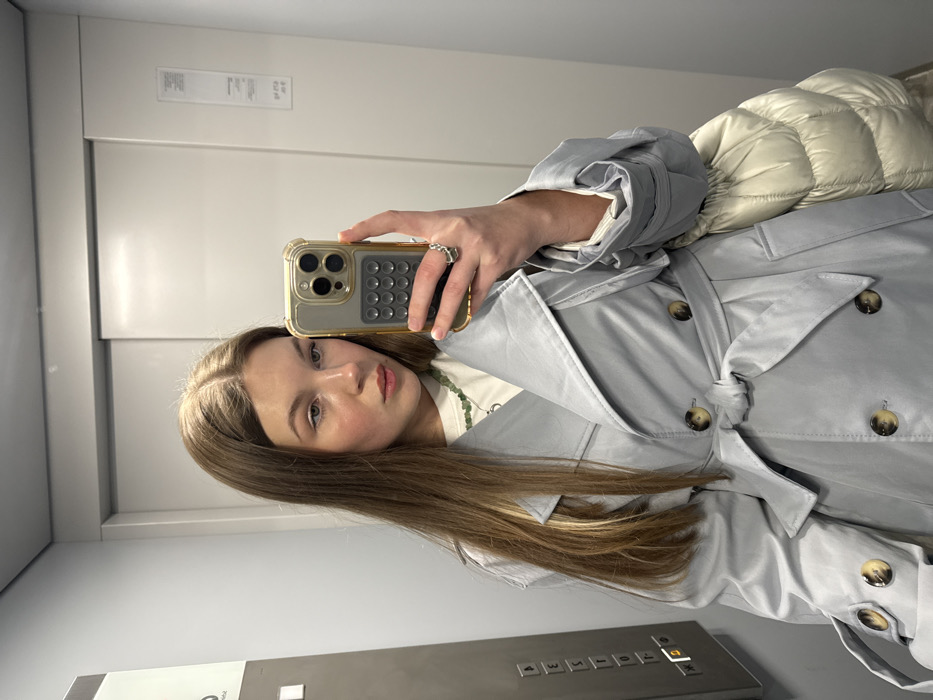 The image displays a young adult female, appearing Caucasian, with long, light brown hair, taking a mirror selfie inside an elevator. She occupies the center-left of the frame, facing forward. She is wearing a light gray double-breasted trench coat with a tied belt and brown buttons, revealing a white t-shirt and a green beaded necklace underneath. Her left hand holds a gold-colored smartphone with a clear case and a dark gray, grid-patterned accessory on the back, aimed at a reflective surface. A silver-toned ring is visible on her left ring finger. A cream-colored quilted garment or bag is slung over her left arm.
The environment is an indoor elevator, characterized by light gray metallic walls. To the left, a control panel with silver buttons and an illuminated up arrow is visible. The reflective surface she uses for the selfie is the polished metal of the closed elevator doors, which feature a horizontal seam and a vertical panel on the right. No specific street address or unique identifying landmarks within the Vozdovac Urban Municipality, Serbia, are discernible from the image to pinpoint the exact location.
