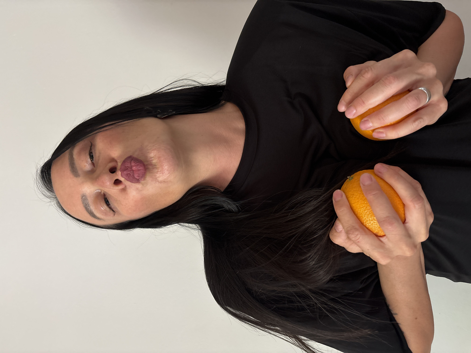 A woman with long, dark hair wearing a black shirt is holding two oranges. She is making a kissing face. The background is a plain white wall. There is no indication of the location being Chaikovsky, Russia.