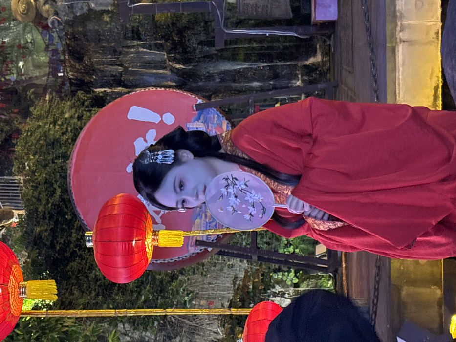 The image displays a young East Asian female in traditional red Hanfu attire, standing in an illuminated outdoor environment within Yuzhong District, Chongqing, China. She is positioned slightly right of the frame's center, her gaze directed subtly downward and to the left. Her dark hair is styled in an intricate bun, adorned with silver-colored, dangling hair ornaments. She holds a white circular fan with a white floral pattern, partially obscuring her mouth and lower face. Two illuminated red Chinese lanterns with yellow tassels are visible on the left, with another red object partially present in the bottom left. Behind the female, a large red circular disc structure features white Chinese characters, "重庆!" (Chóngqìng!), above a landscape painting. The background includes dark green foliage, an illuminated rocky waterfall to the right of the disc, and a dark architectural structure resembling a bell stand on the far right. The ground in the immediate foreground is paved.
