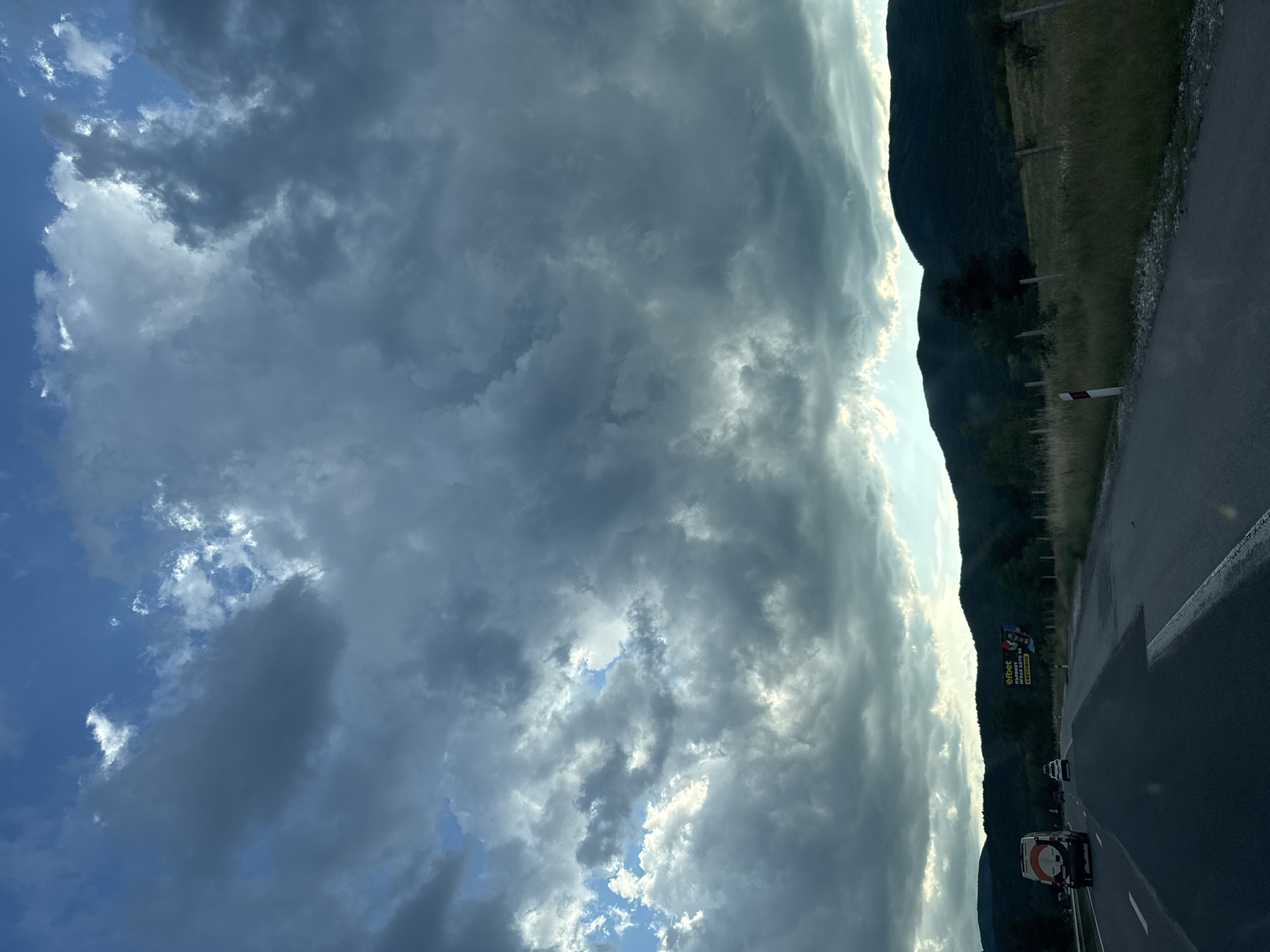 The image depicts a view from a vehicle traveling on a paved road, likely a highway, heading towards a landscape with rolling hills and mountains in the background. The sky is dramatic, filled with dark, heavy clouds interspersed with patches of bright sunlight breaking through. The road itself has clear lane markings, and there are other vehicles on it, including a white van and a smaller white car further ahead. To the right of the road, there is a grassy verge with a wire fence and wooden posts. A large billboard advertising "efbet" is visible on the right side of the road, featuring colorful imagery and Bulgarian text. The overall impression is of a journey through a rural or semi-rural area under a dynamic, possibly approaching weather system. The location is identified as Gorno Varshilo, Bulgaria.