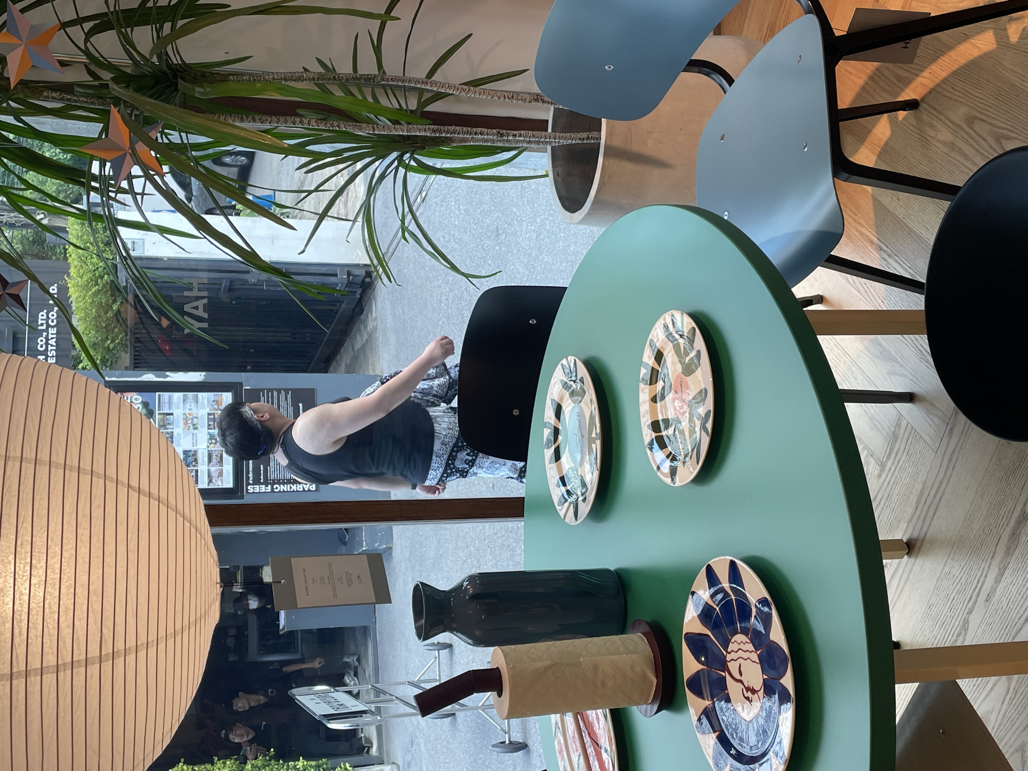 The image depicts an interior scene with a view to the outdoors. There are people visible inside, some seated at tables and others standing. A person wearing a black tank top and patterned pants appears to be walking. A round green table adorned with decorative plates is in the foreground, and there are blue and black chairs around it. On the table, a dark green vase and a roll of paper towels on a holder are visible. A large, round paper lamp is partially visible on the left side.
The outdoor view shows a street scene with vehicles and greenery. A sign mentions "PARKING FEES" and "A CO., LTD. ESTATE CO., D.". A store sign reads "YAH".
Based on the OCR text, it can be concluded that the location is in Bangkok, Thailand, and the scene shows part of a commercial establishment.