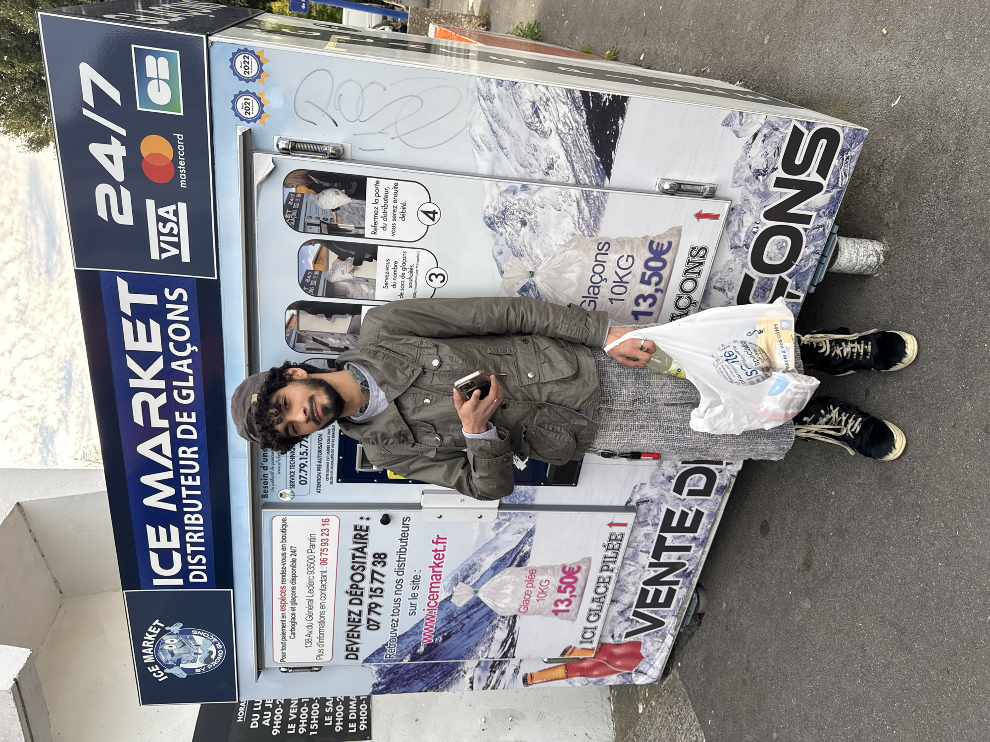 A man stands in front of an ice vending machine. The vending machine is located outdoors on a street, possibly in Pantin, France, according to the text on the machine. The machine is white and blue, with logos for Visa and Mastercard. It advertises ice for sale 24/7. The man is holding a cell phone in his left hand and a plastic bag full of various items in his right hand. He is wearing a khaki jacket, gray pants, and black shoes. The vending machine displays prices for ice: Glace pilée 10KG, 13.50€. There are also instructions for using the machine. The URL www.icemarket.fr is printed on the machine.