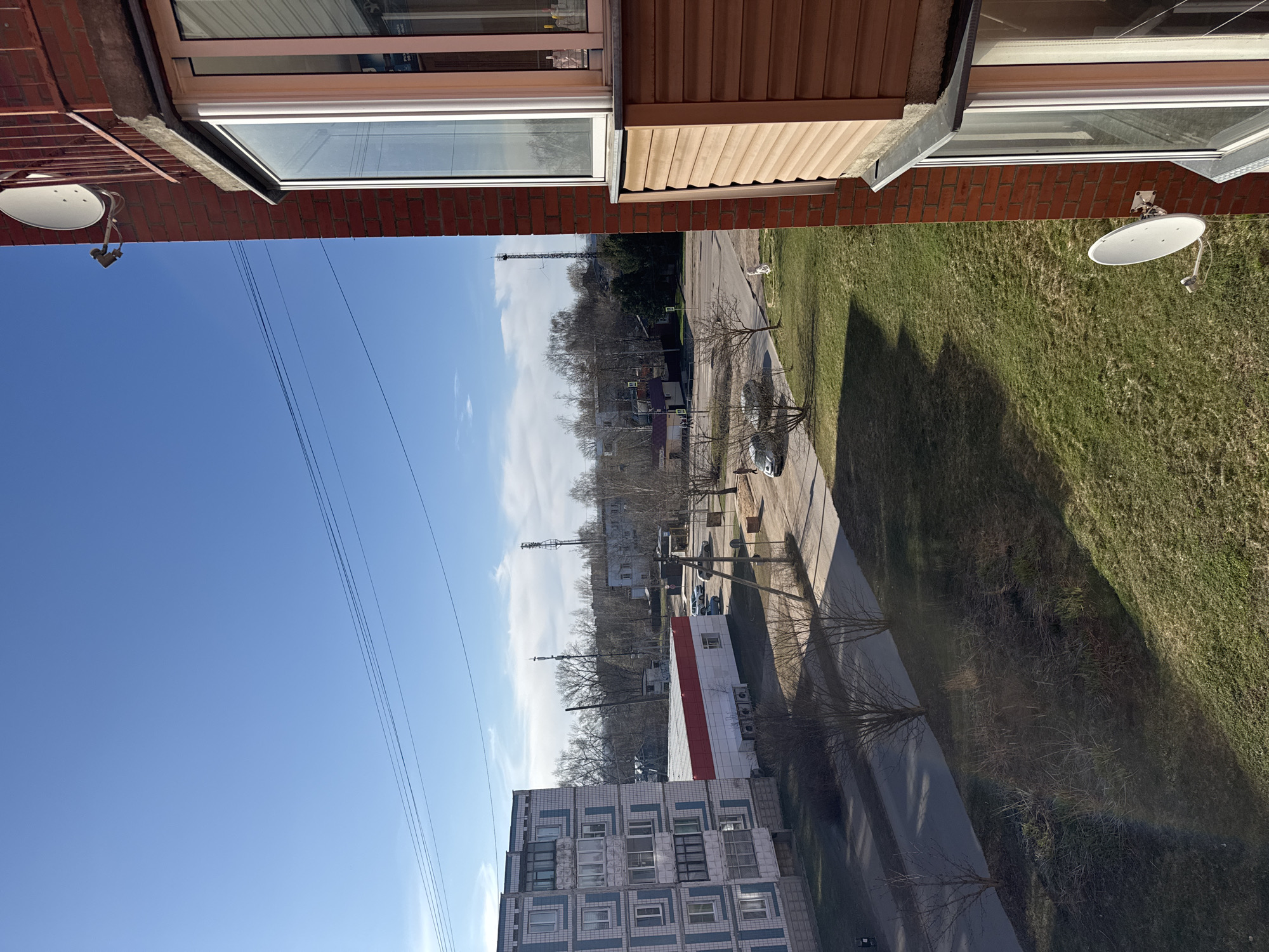 The image captures a view from an elevated position, likely a high-rise building, in Dmitrov, Russia. Below, one can see a typical urban landscape.
**Setting:** The scene showcases a clear blue sky with some scattered clouds. In the foreground, there is the red brick side of a building, presumably where the photo was taken, including a white satellite dish affixed to it.
**Objects:** A road is visible that is lined with trees and a grassy area. Some buildings are shown in the distance. Two communication towers can also be identified in the distance. The architectural style of the buildings suggests a post-Soviet design, typical for many Russian cities. A red-and-white commercial building is also visible.
**People:** The scene does not contain any discernible individuals.
**Actions:** There are no actions captured in the scene.
**Interactions:** No interactions can be seen.
**Environment:** The image gives an impression of a day in a quiet neighborhood in Dmitrov. The presence of trees and green areas suggests a relatively green urban environment.